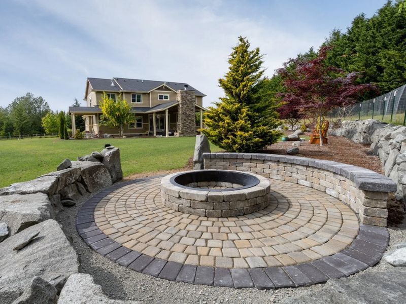 Circular stone fire pit in a landscaped backyard patio with a house and lawn in the background