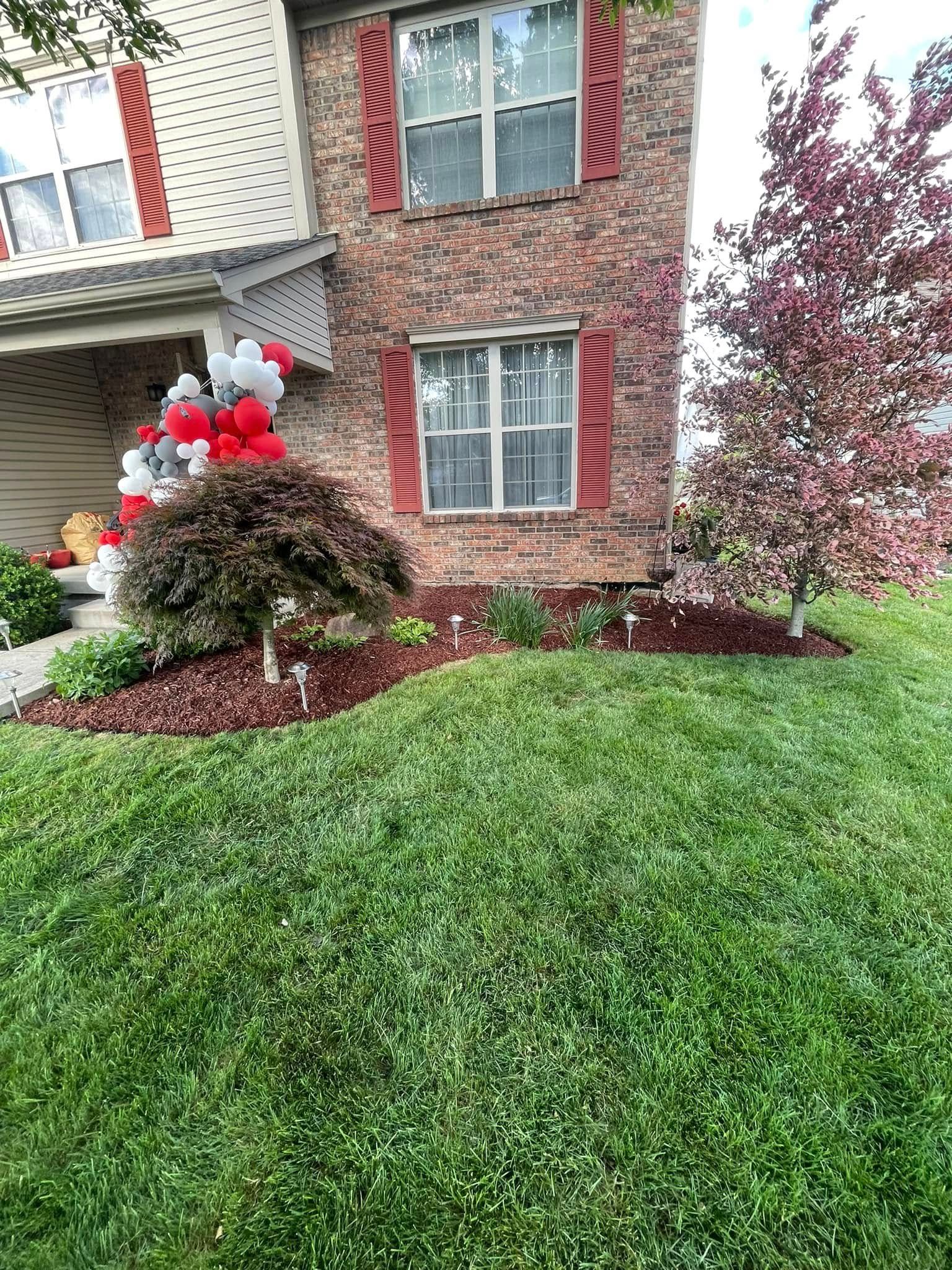 Front yard of a brick house with a landscaped bed, small tree, and red-and-white balloon decorations near the porch.