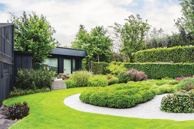 Lush landscaped garden with curved white path, green shrubs, trees, and a small modern black shed