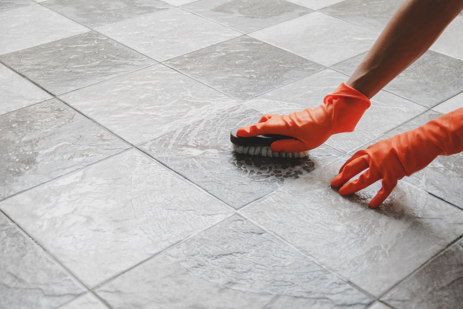 Hands in orange gloves scrubbing a gray tiled floor with a brush.