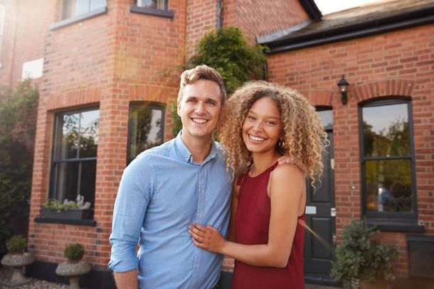 A smiling couple stands arm-in-arm in front of a red brick house during the day.