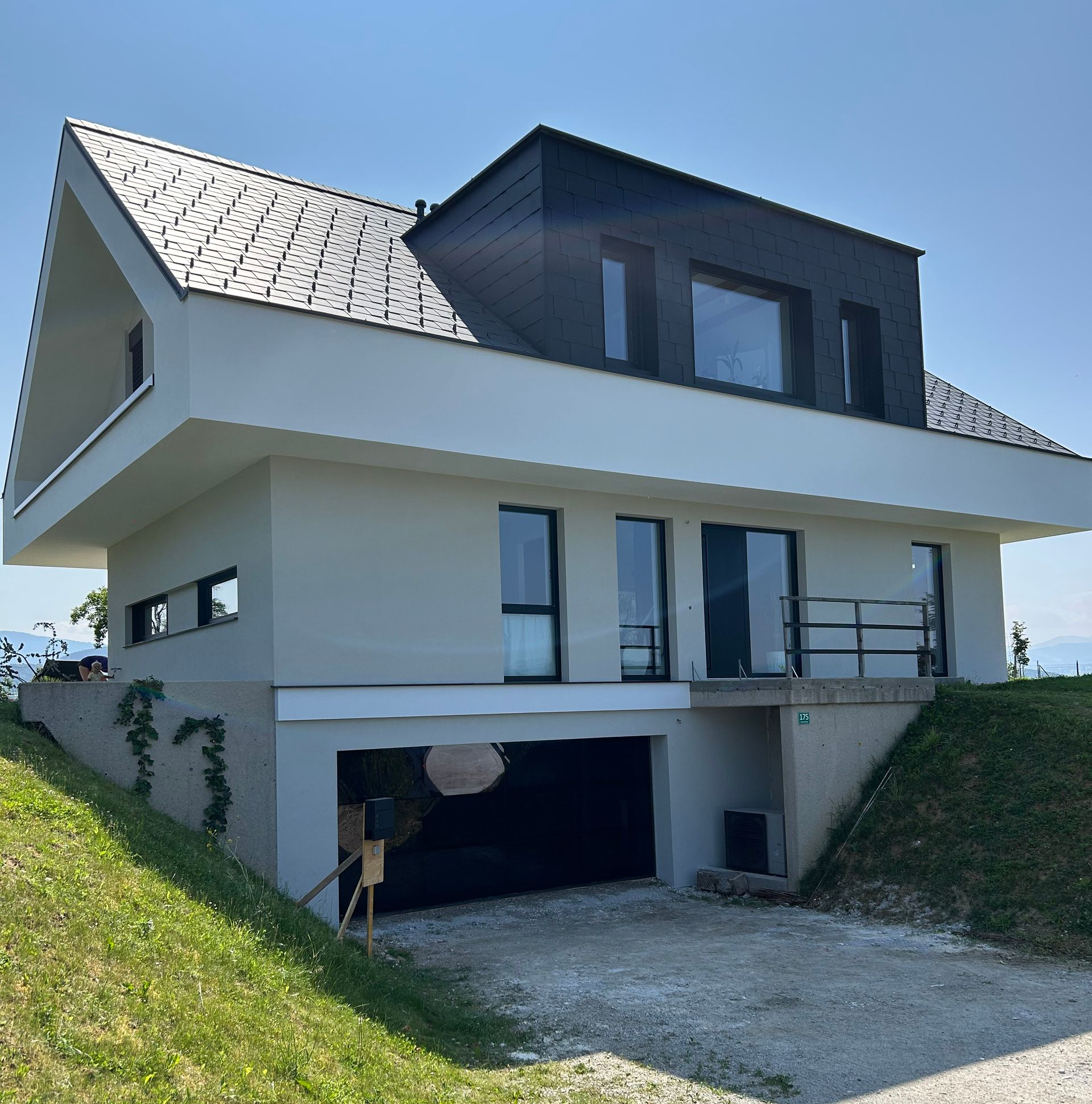 Modernes, weißes zweistöckiges Haus mit dunklem Dach, Balkon, Garage und abfallender grüner Rasen unter klarem, blauem Himmel