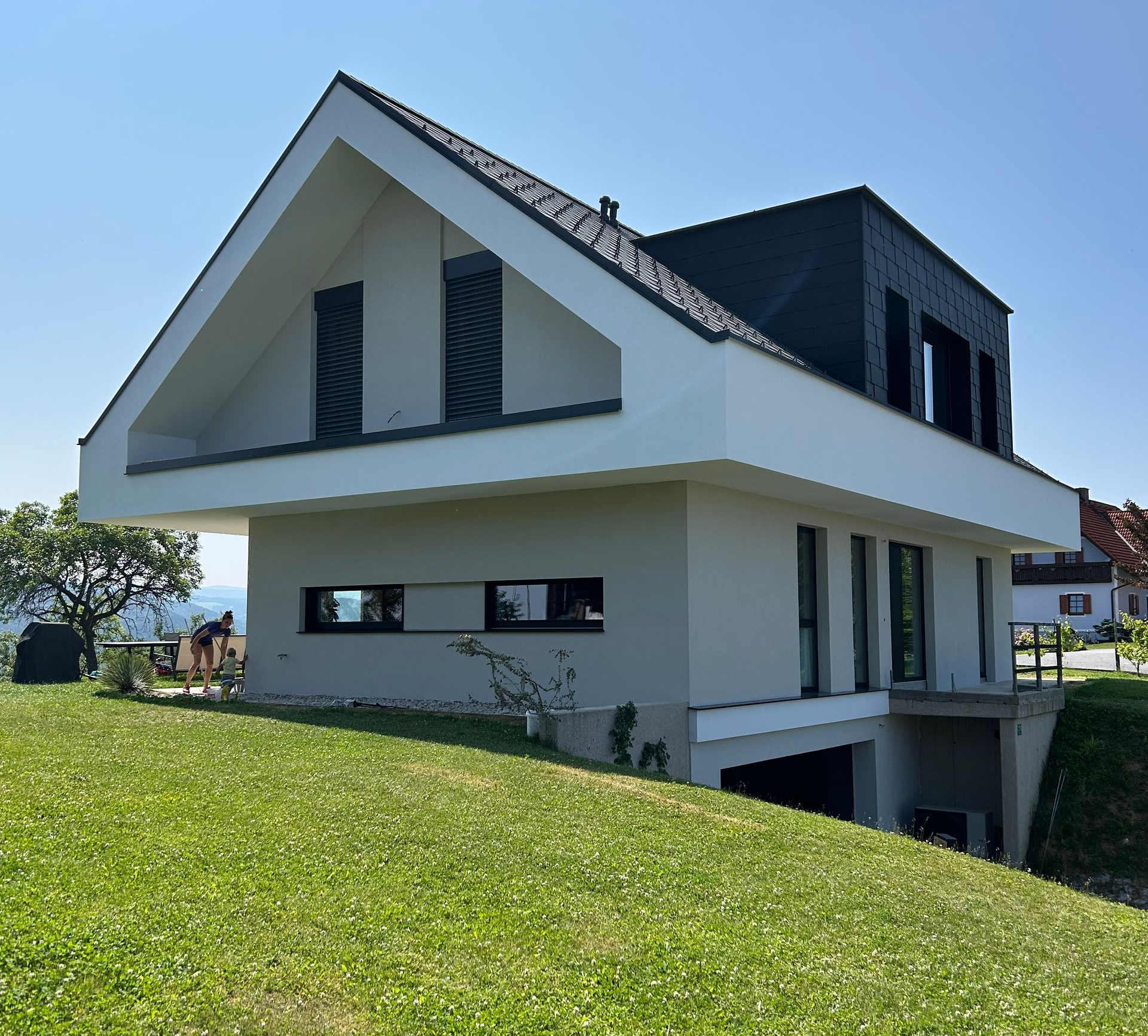Modernes weißes Haus mit kantigem Dach an einem grasbewachsenen Hang unter klarem blauen Himmel