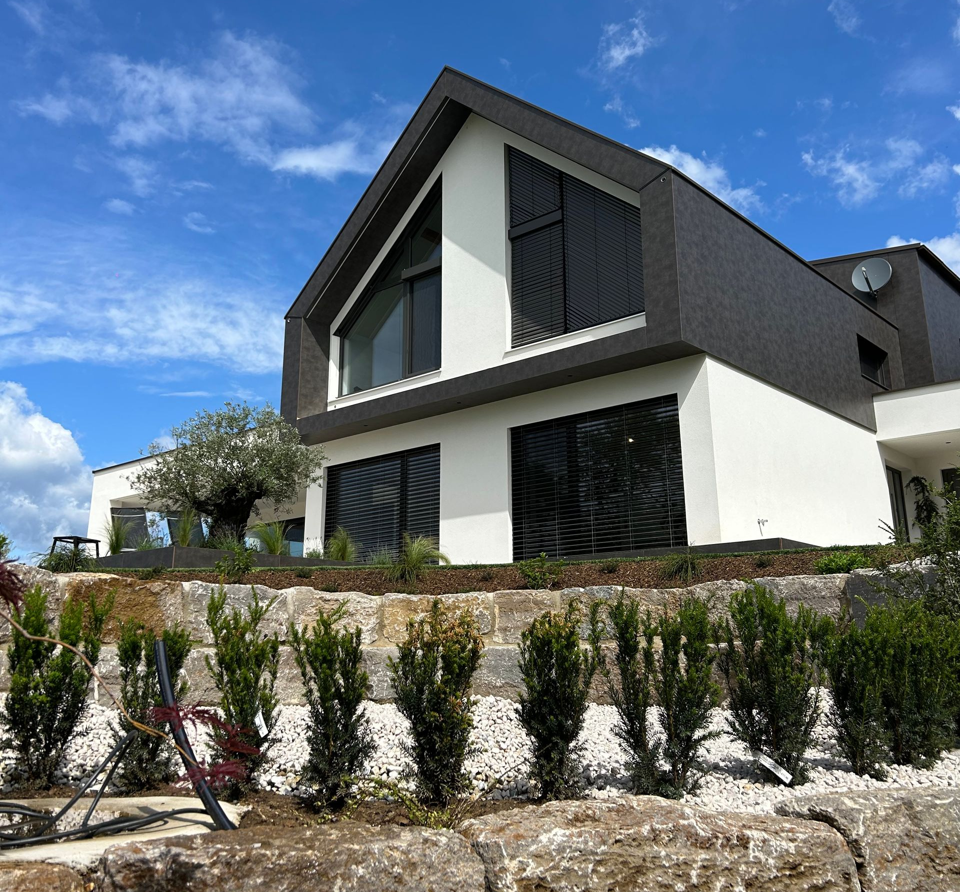 Modernes zweistöckiges Haus mit dunklem Dach, weißen Wänden und einer landschaftlich gestalteten Steinfassade