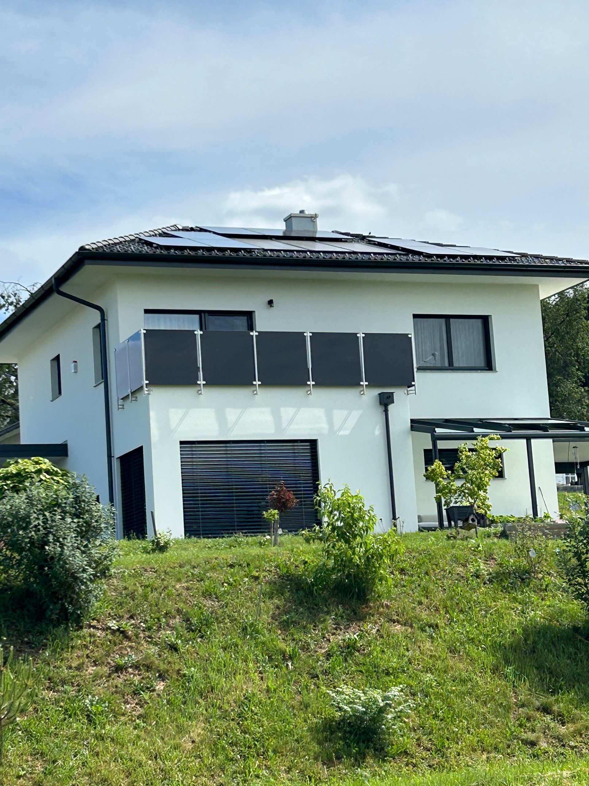 Modernes weißes zweistöckiges Haus mit Solarpaneelen auf dem Dach, gesehen von einem grasbewachsenen Hang unter blauem Himmel
