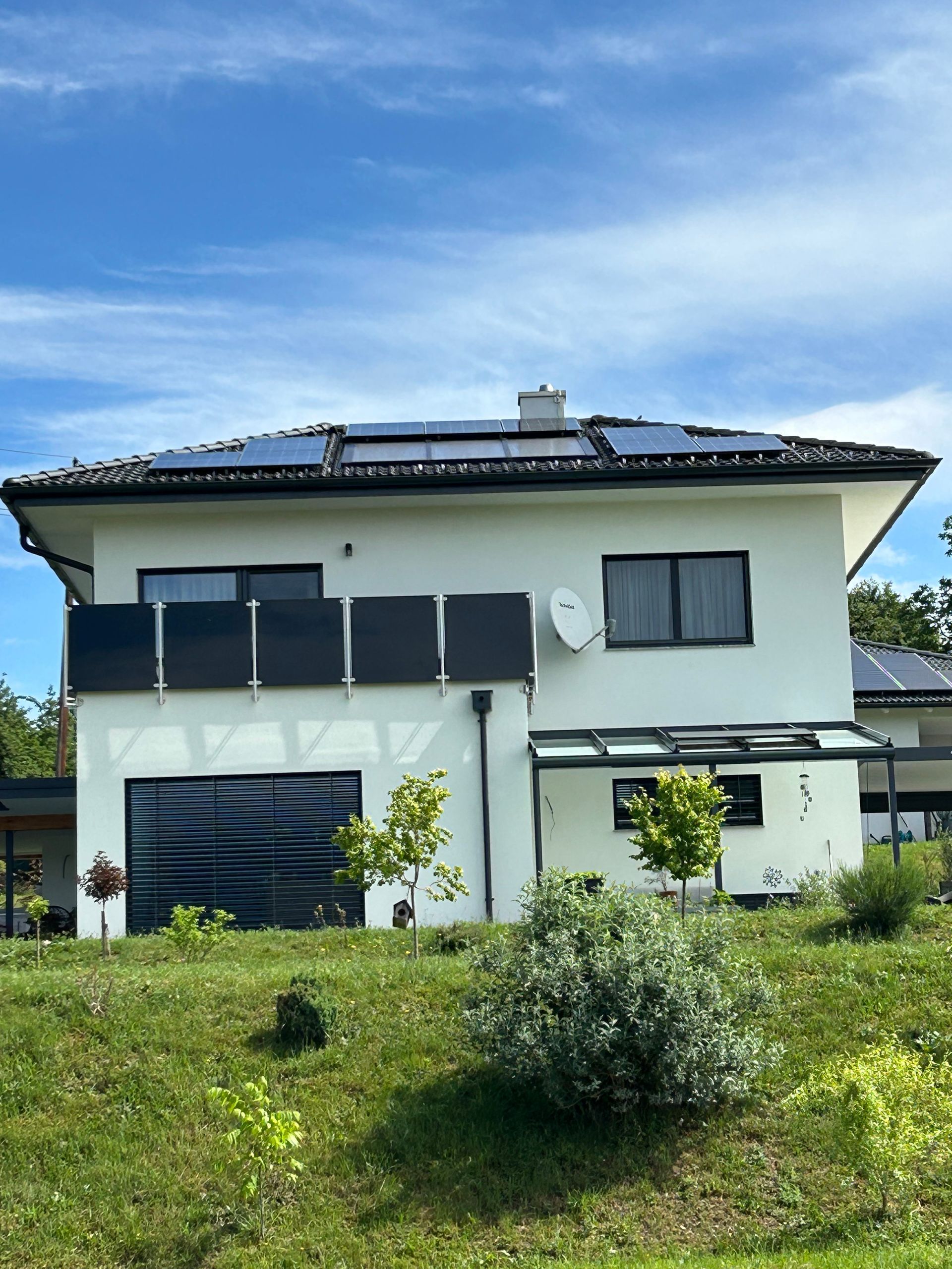 Zweistöckiges weißes Haus mit dunklem Dach, Balkon und grünem Rasen unter blauem Himmel