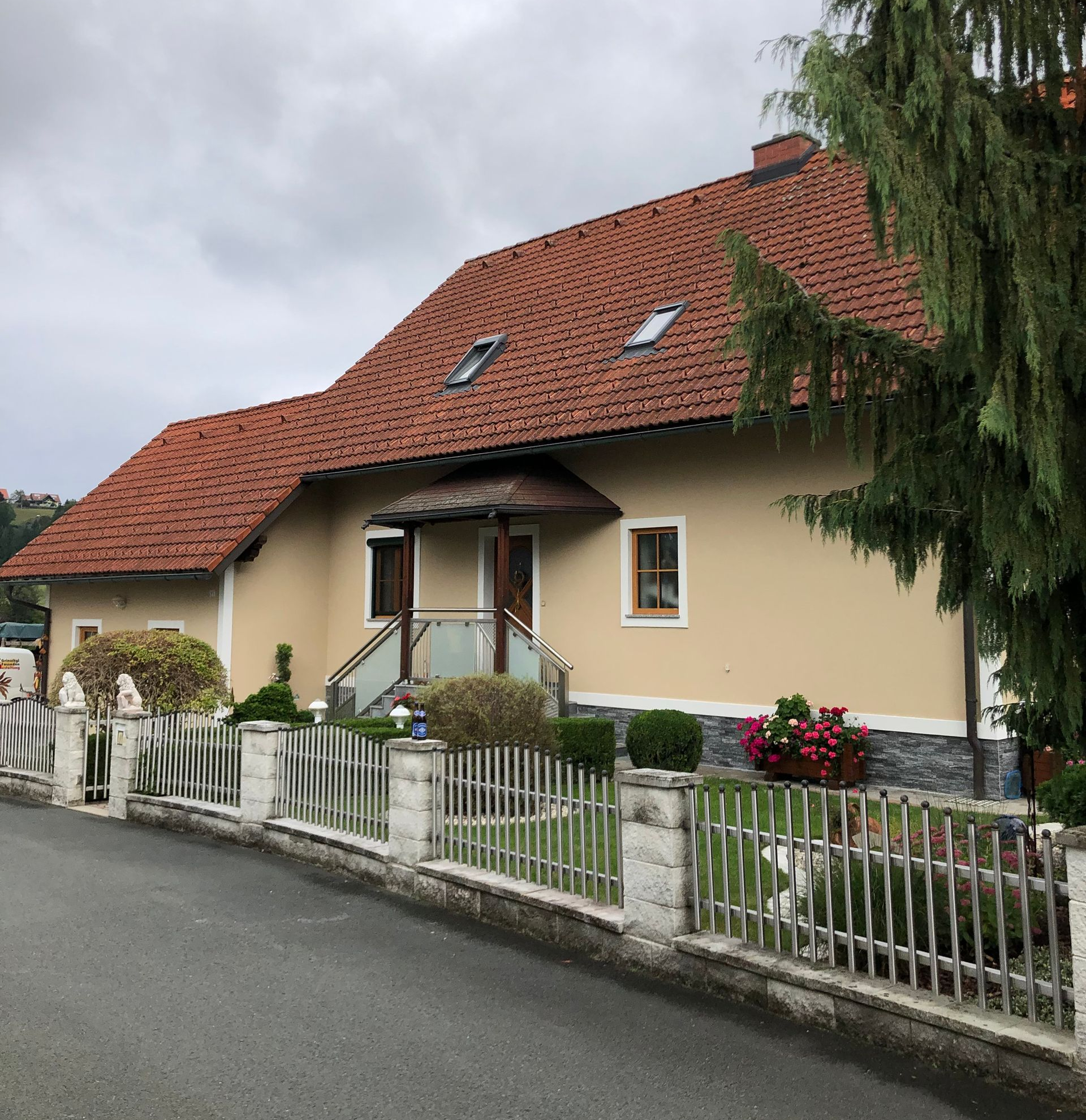 Haus mit rotem Ziegeldach und weißem Zaun in einer ruhigen Straße unter einem bewölkten Himmel