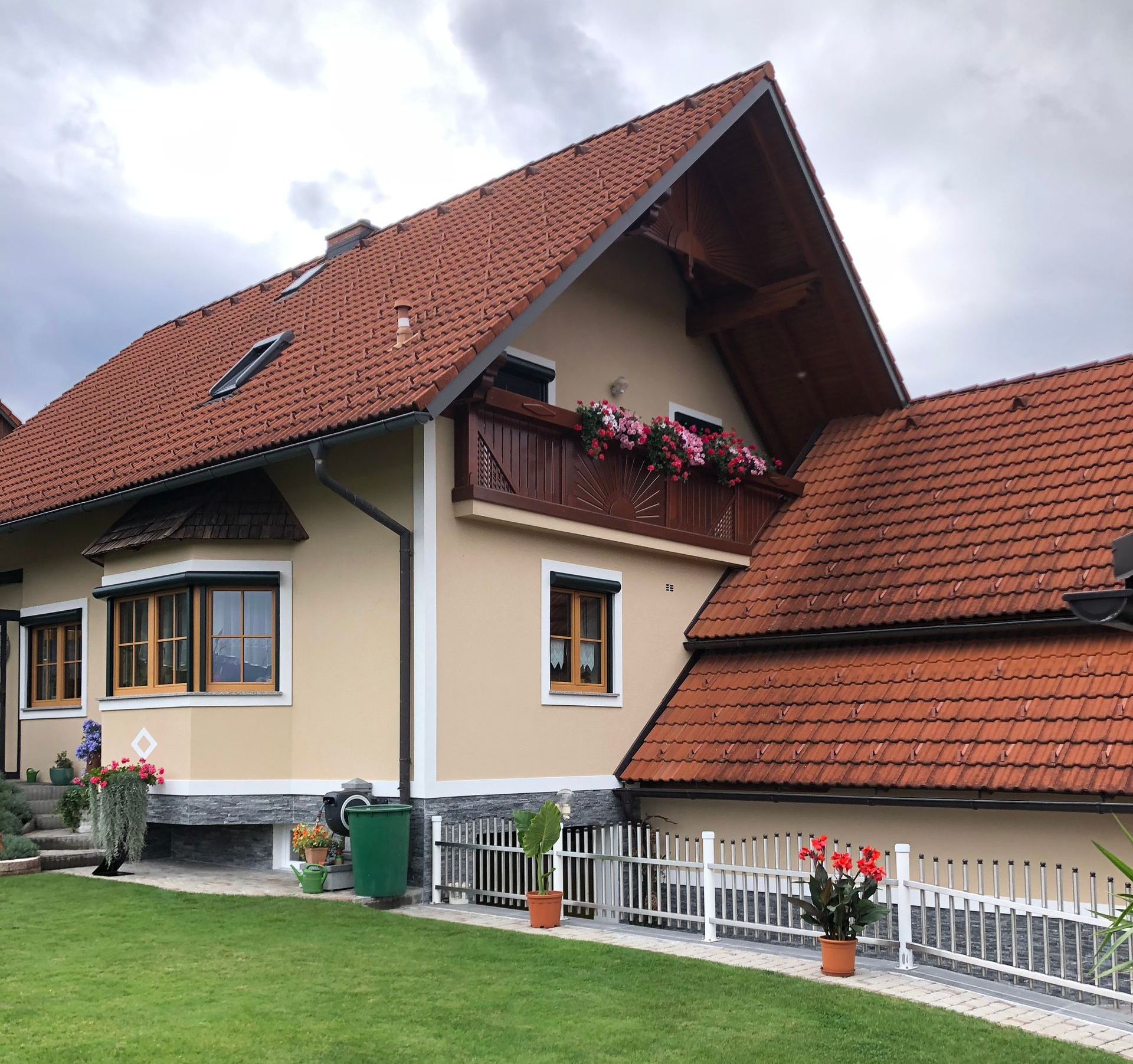 Haus mit rotem Ziegeldach, cremefarbenen Wänden, Blumenkästen und einem grünen Rasen unter einem bewölkten Himmel