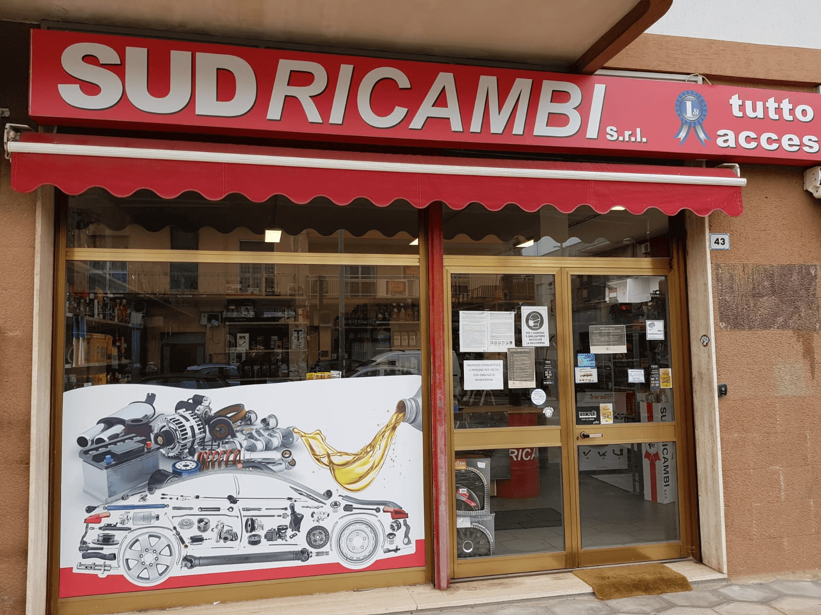 Vetrina del negozio “SUDRICAMBI” con tenda da sole rossa e ricambi auto esposti in vetrina.