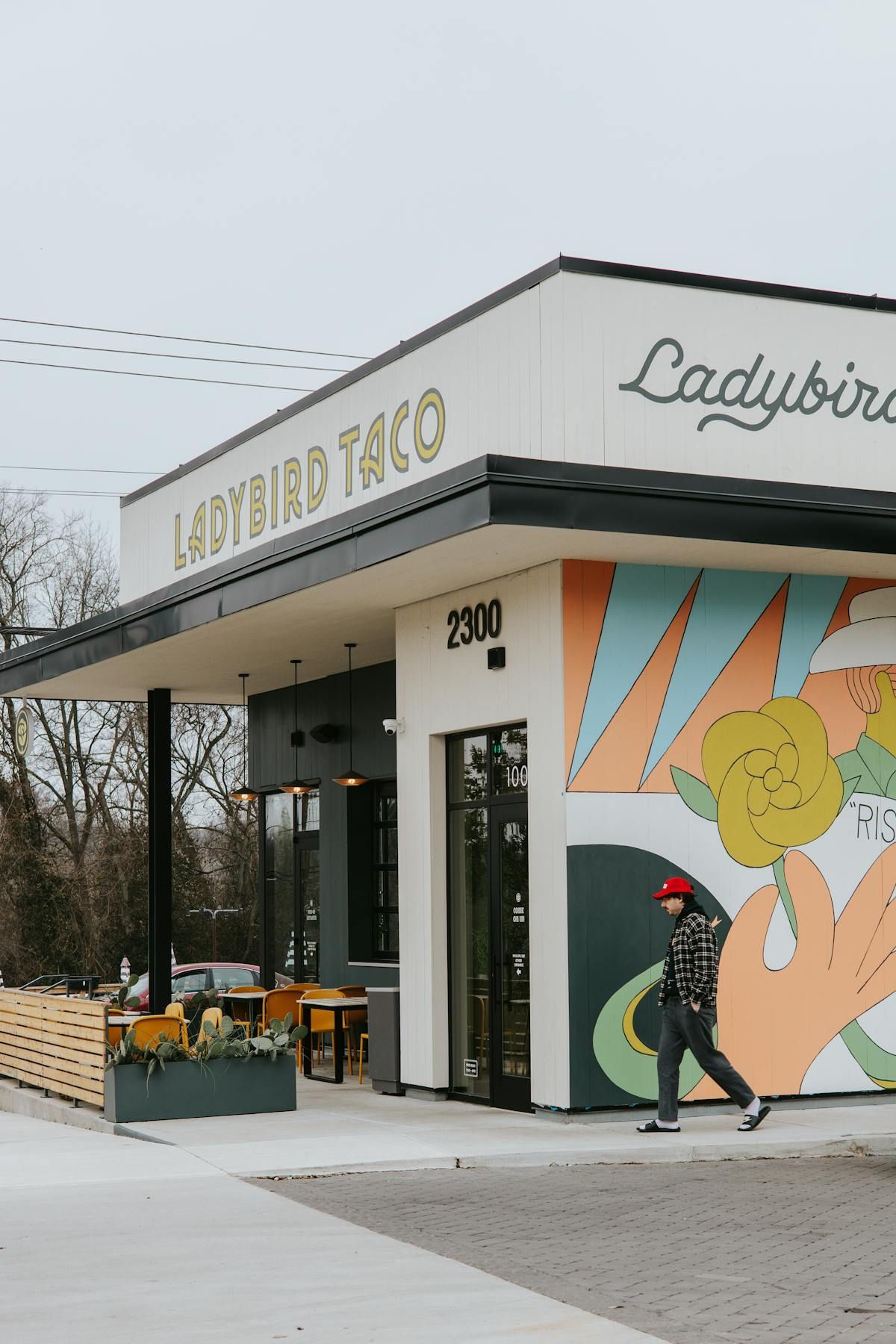 A person walks past Ladybird Taco, a cafe with a painted floral mural on its exterior and outdoor patio seating.