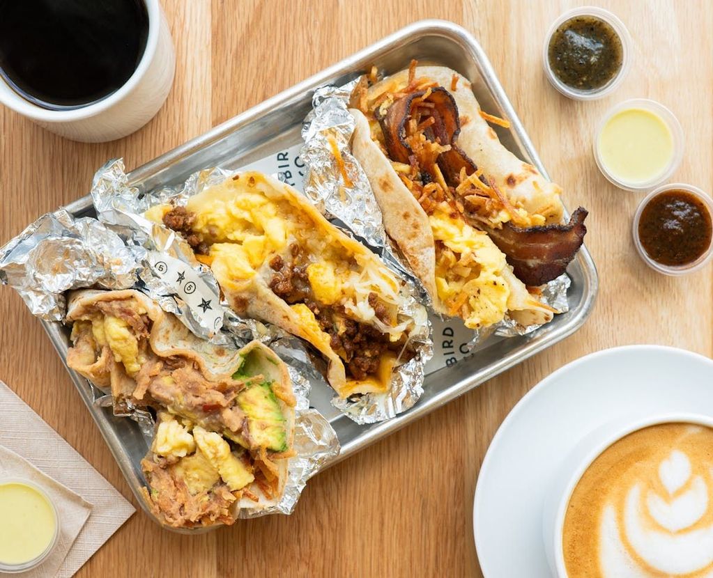 Three breakfast tacos on a metal tray with a cup of coffee, a latte, and three small containers of dipping sauces.