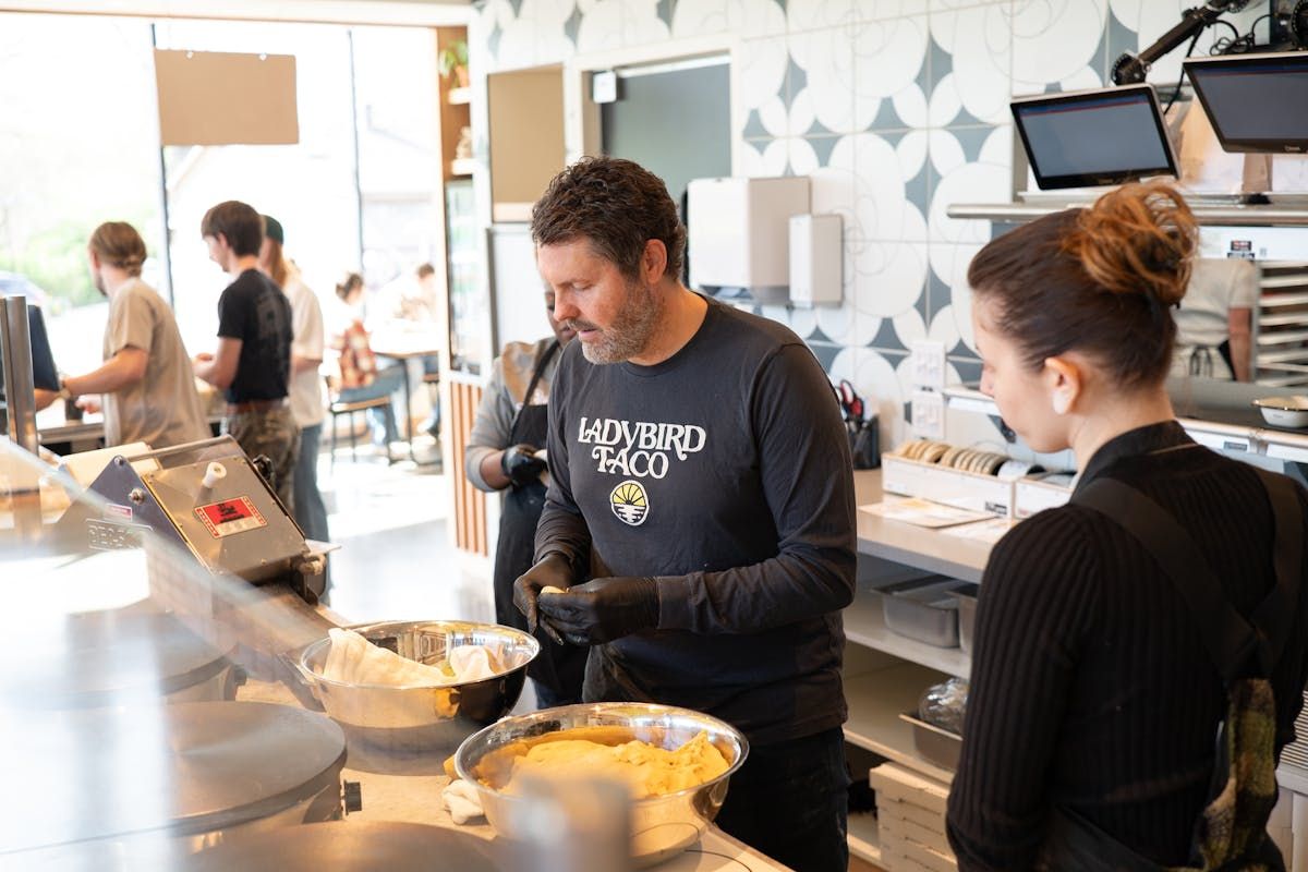 A chef in a black long-sleeve shirt and gloves works behind a kitchen counter, preparing food for a customer.