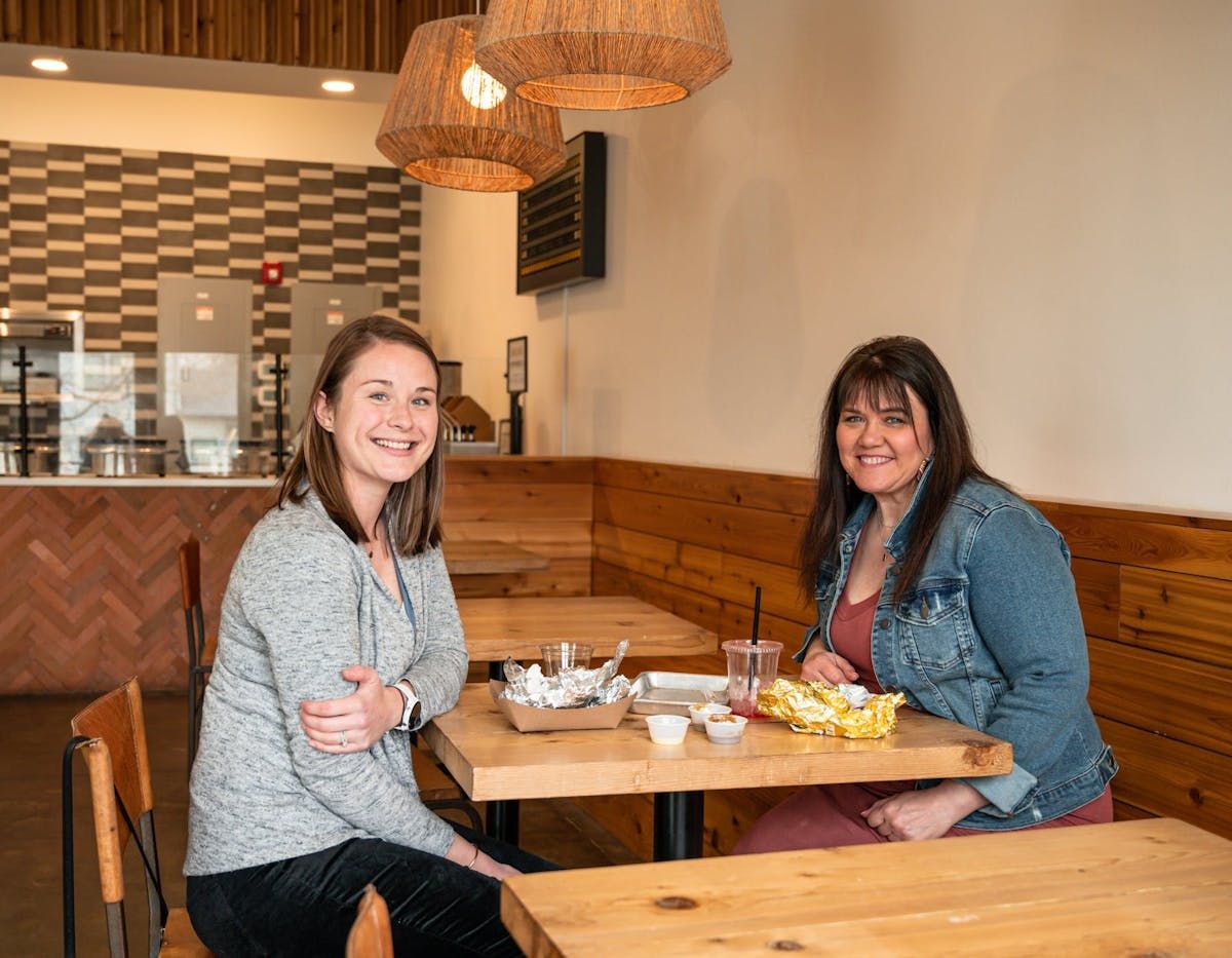 Two people sit at wooden tables in a cafe with textured light fixtures, smiling at the camera while eating a meal.