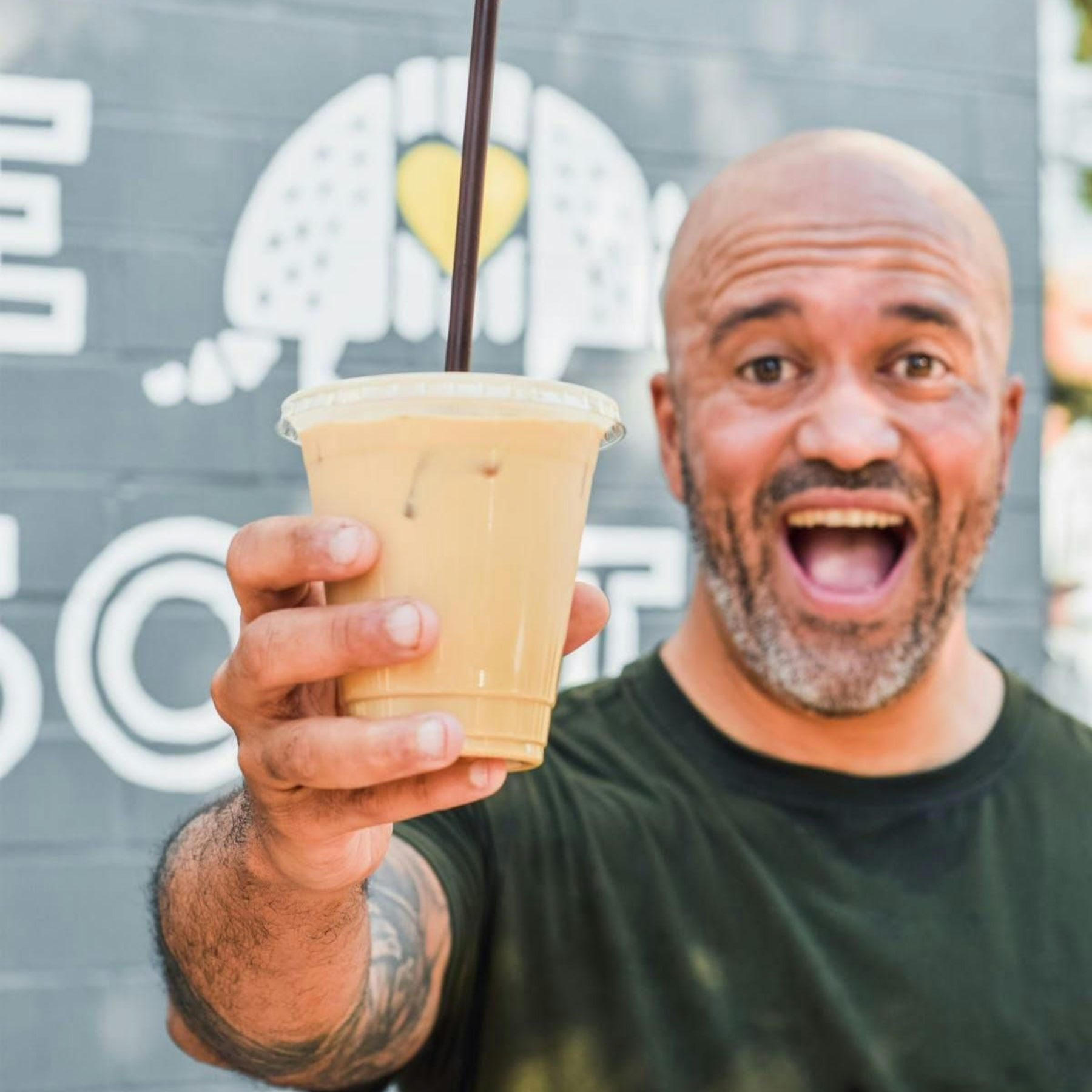 A smiling person holding an iced coffee in a plastic cup with a straw in front of a gray brick wall with an armadillo logo.
