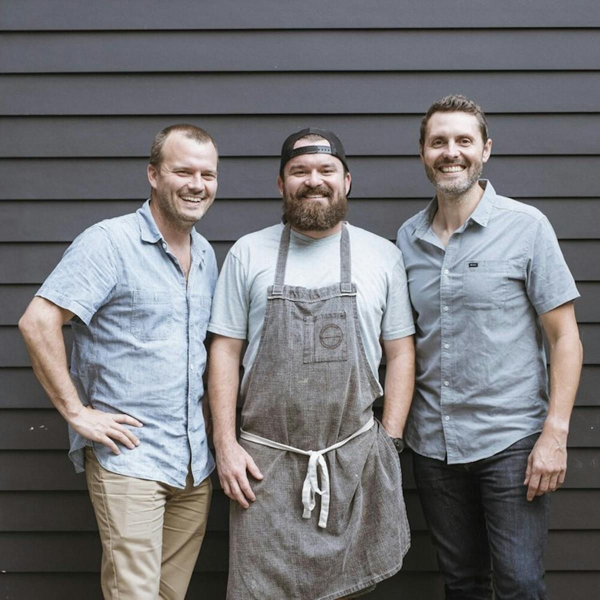 Three smiling people posing side by side against a dark wood-paneled wall, the center person wearing a gray apron.