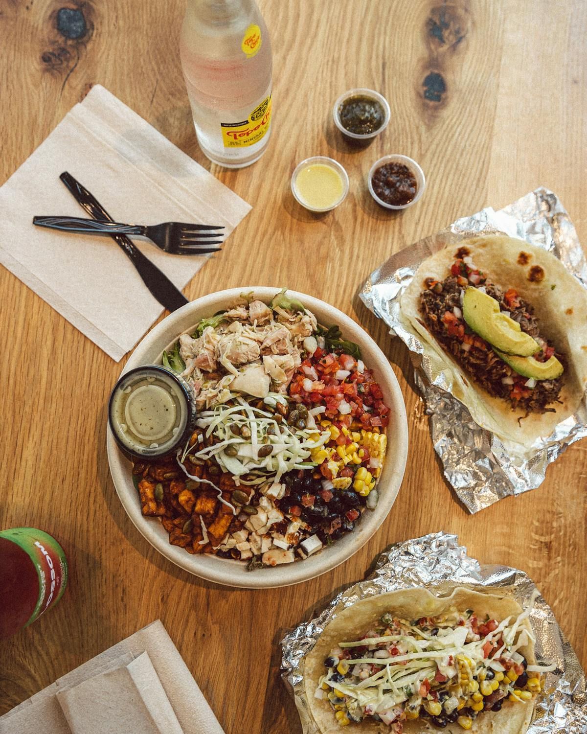 A bowl with chicken and vegetables, two tacos on foil, three small sauce containers, and a drink on a wooden table.