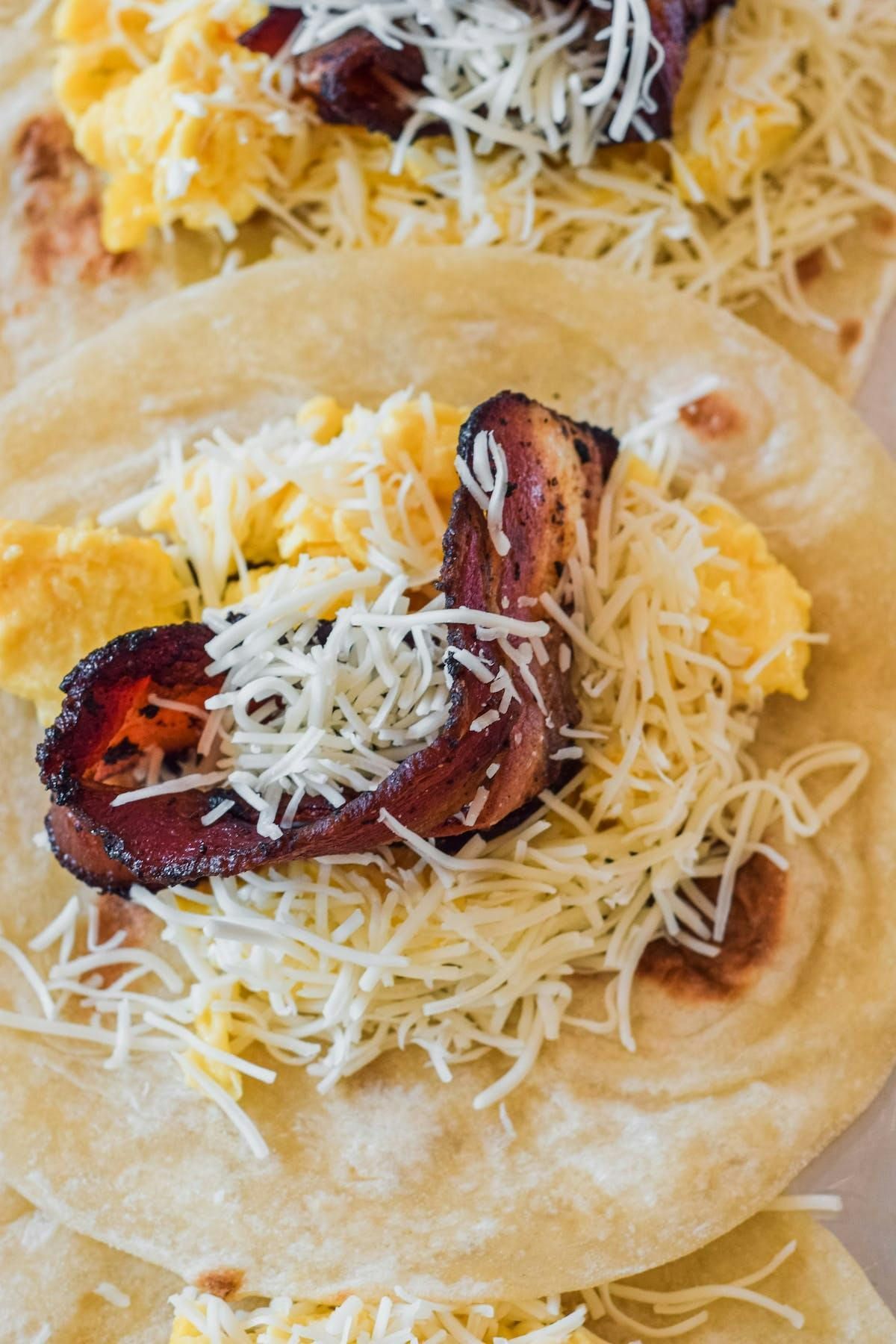 Three breakfast tacos filled with scrambled eggs, crispy bacon, and shredded cheese on flour tortillas.