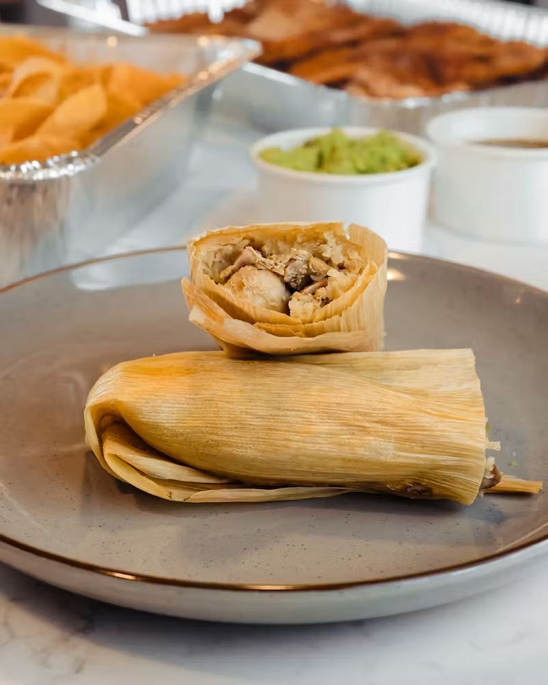 Plate with two chicken tamales, one opened to show filling, on a buffet table with sides in the background