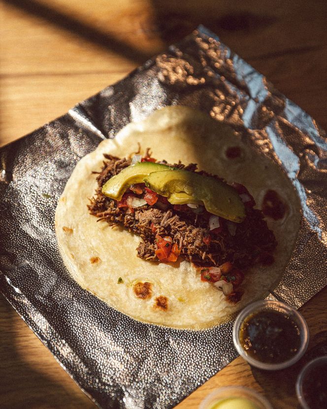 Three breakfast tacos on a metal tray with a cup of coffee, a latte, and three small containers of dipping sauces.