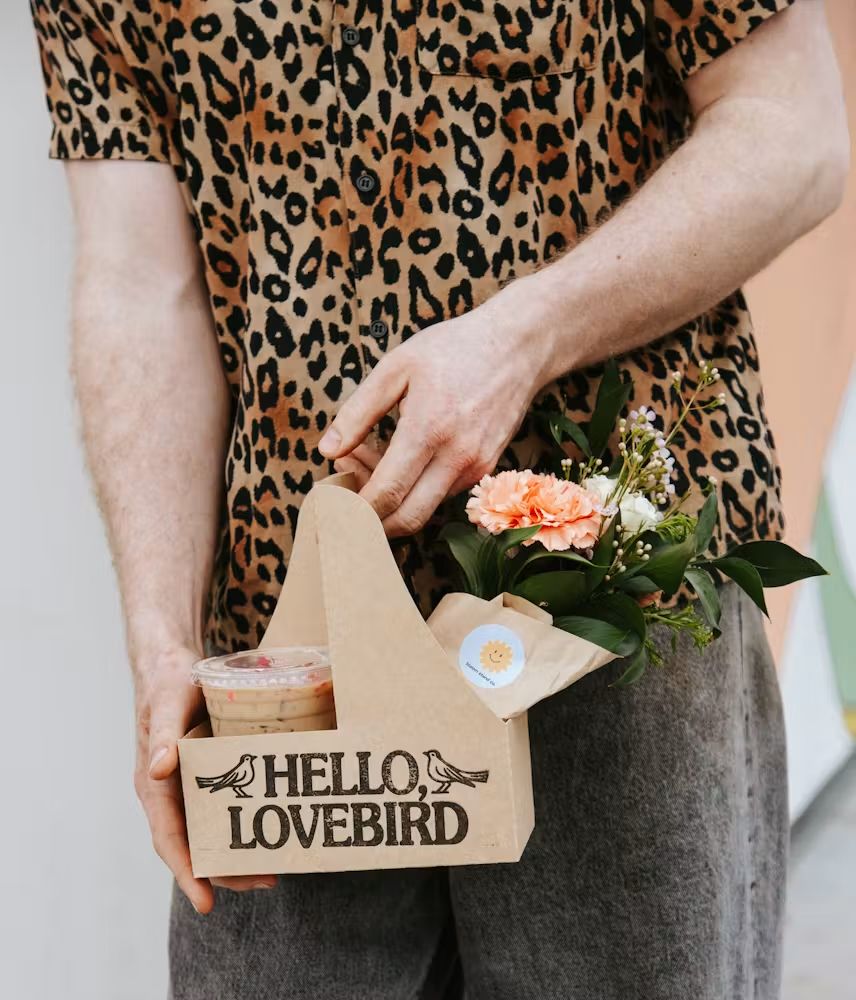 Person holding a small bouquet and an iced coffee in drink carrier labeled “HELLO LOVE BIRD” with two bird illustrations