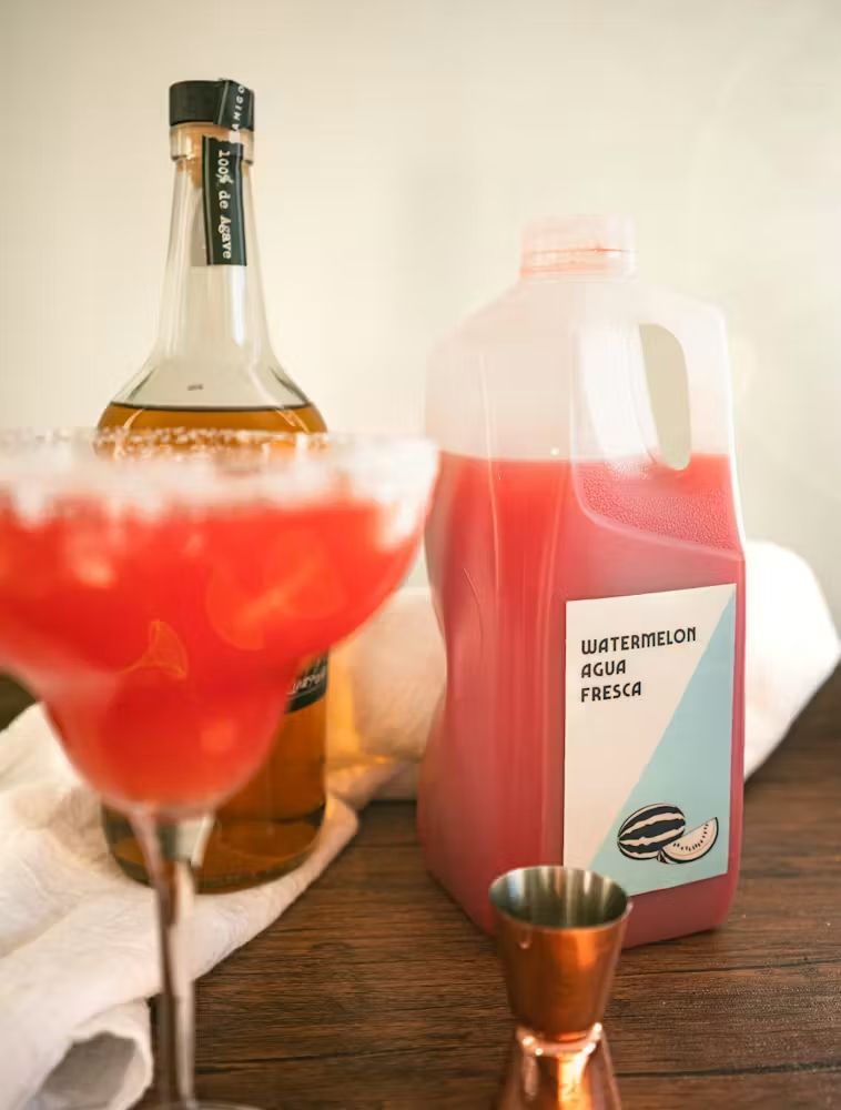 Pink margarita mix beside a liquor bottle and a red drink jug on a wooden table