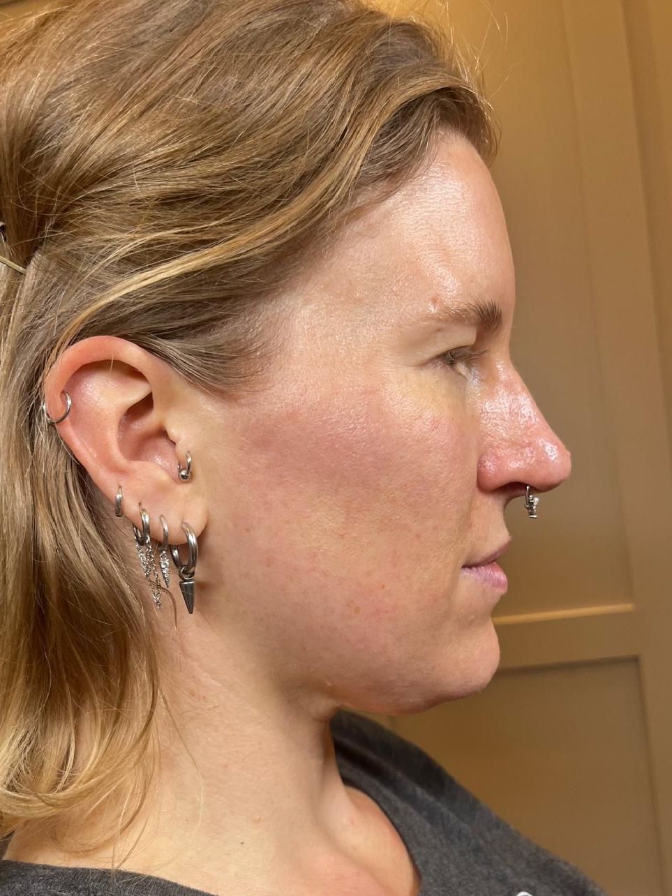 Side profile of a person with blonde hair, multiple earrings, and a nose ring against a warm indoor background