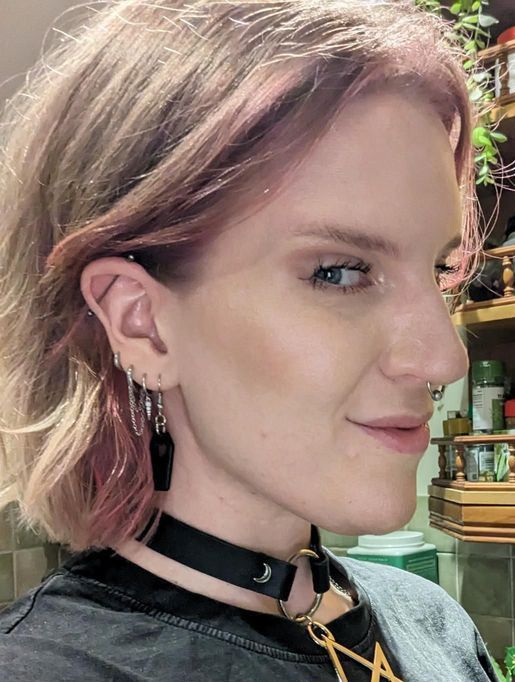 Profile portrait of a person with short pink-blonde hair, black choker, and earrings in a shop setting