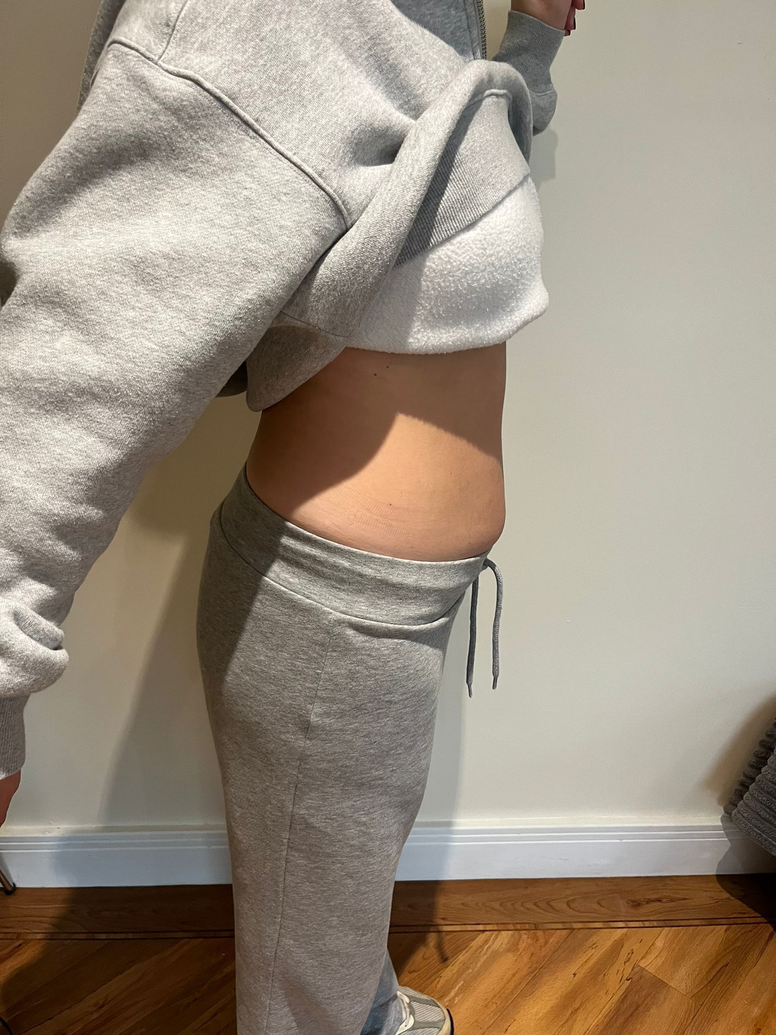 Side view of a person in gray sweats lifting their sweatshirt, showing their waist indoors.