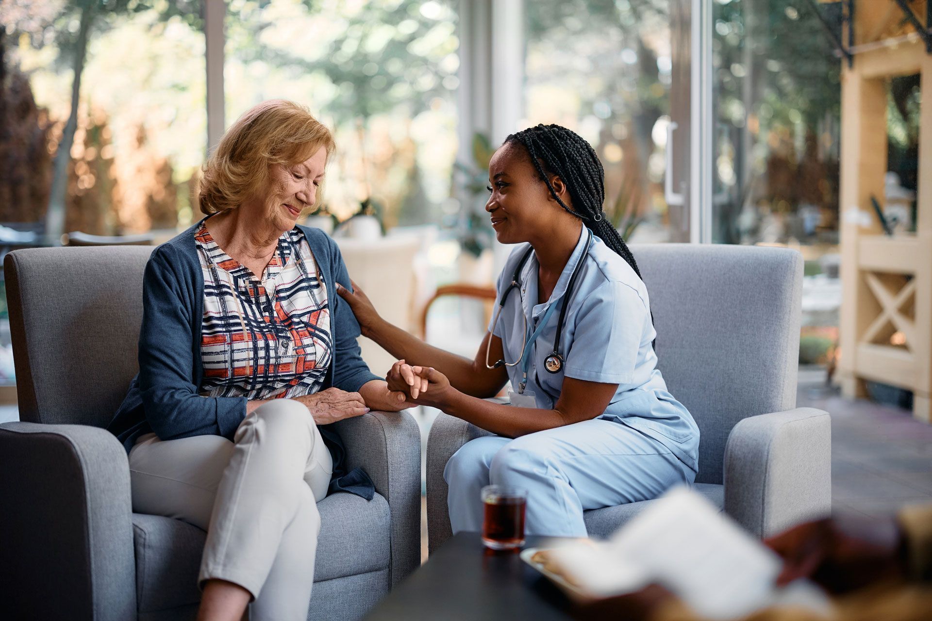 A nurse is holding the hand of an elderly woman while sitting in a chair.
