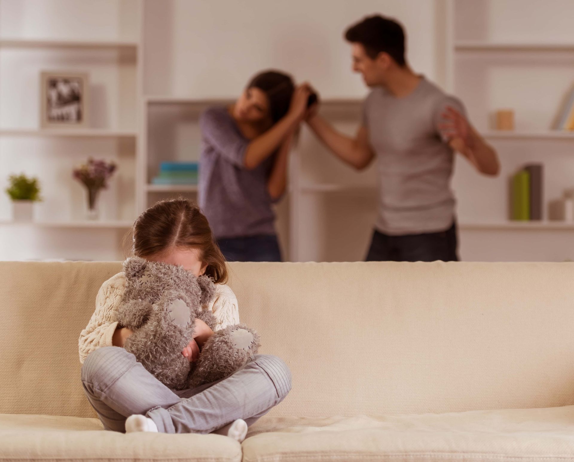 Child sitting curled on a couch, holding a teddy bear, while adults argue in the background