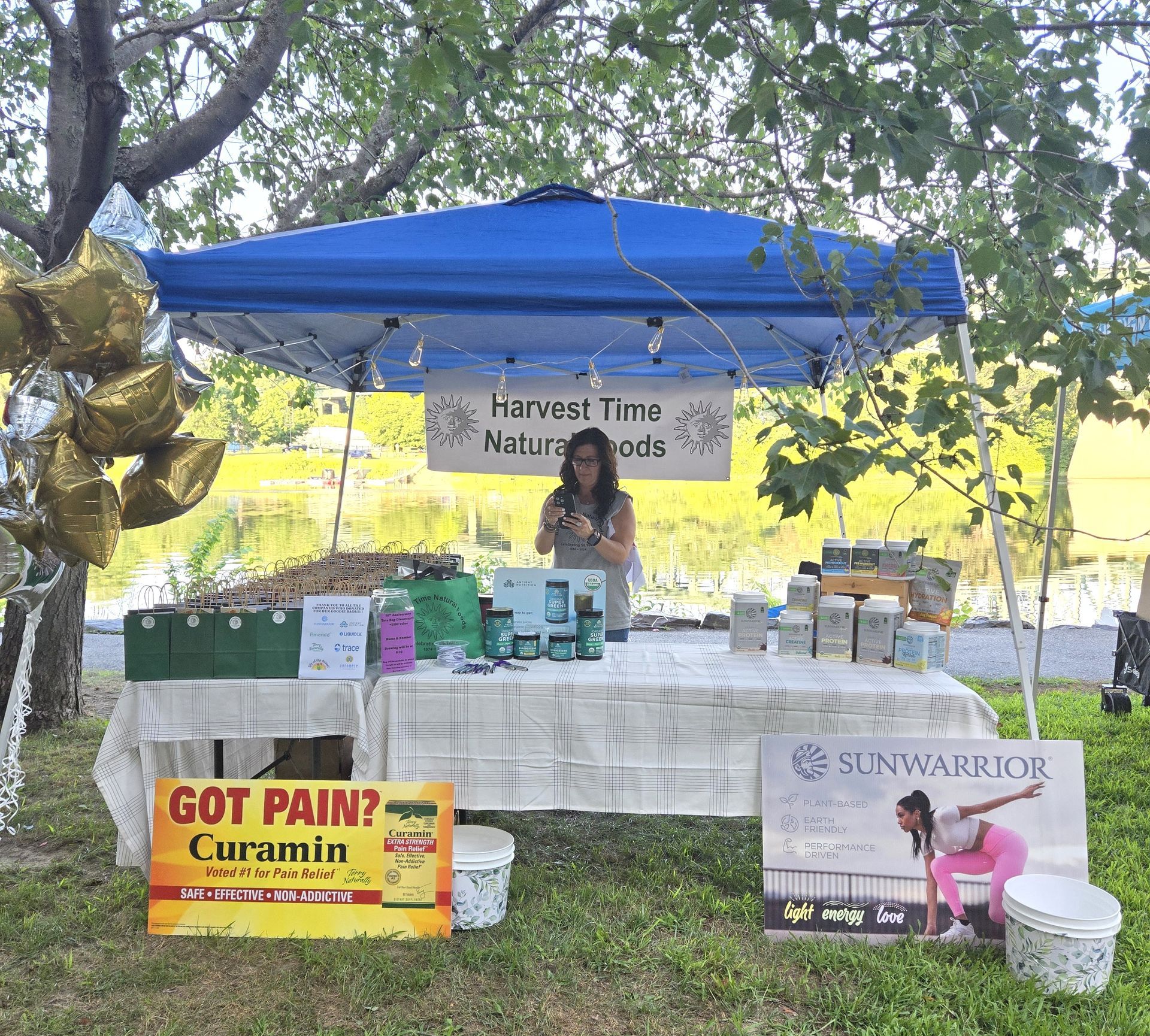 A woman stands behind a Harvest Time Natural Foods stall under a blue canopy, featuring product displays and signs.