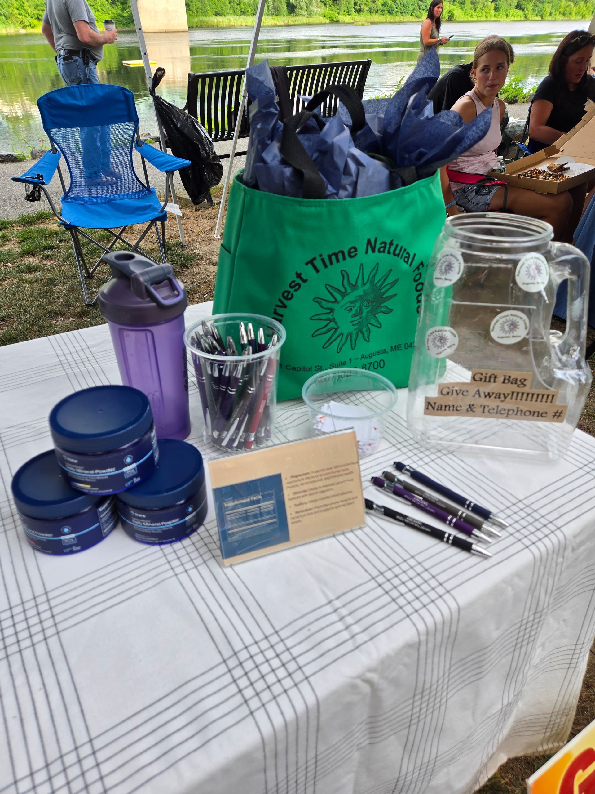 A table at an outdoor event displays a green branded tote bag, a purple shaker cup, pens, and promotional containers.