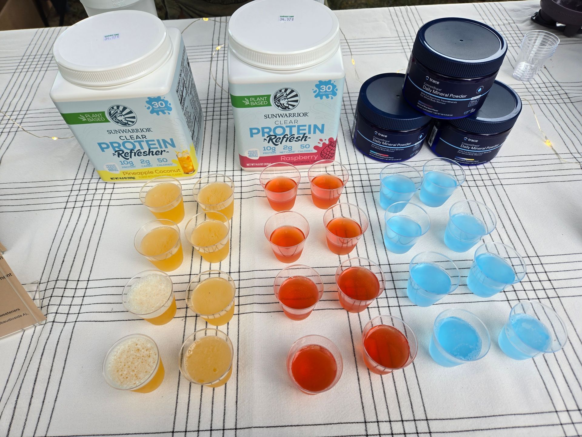 Two large protein powder containers and three supplement jars behind rows of orange, red, and blue liquid sample cups.