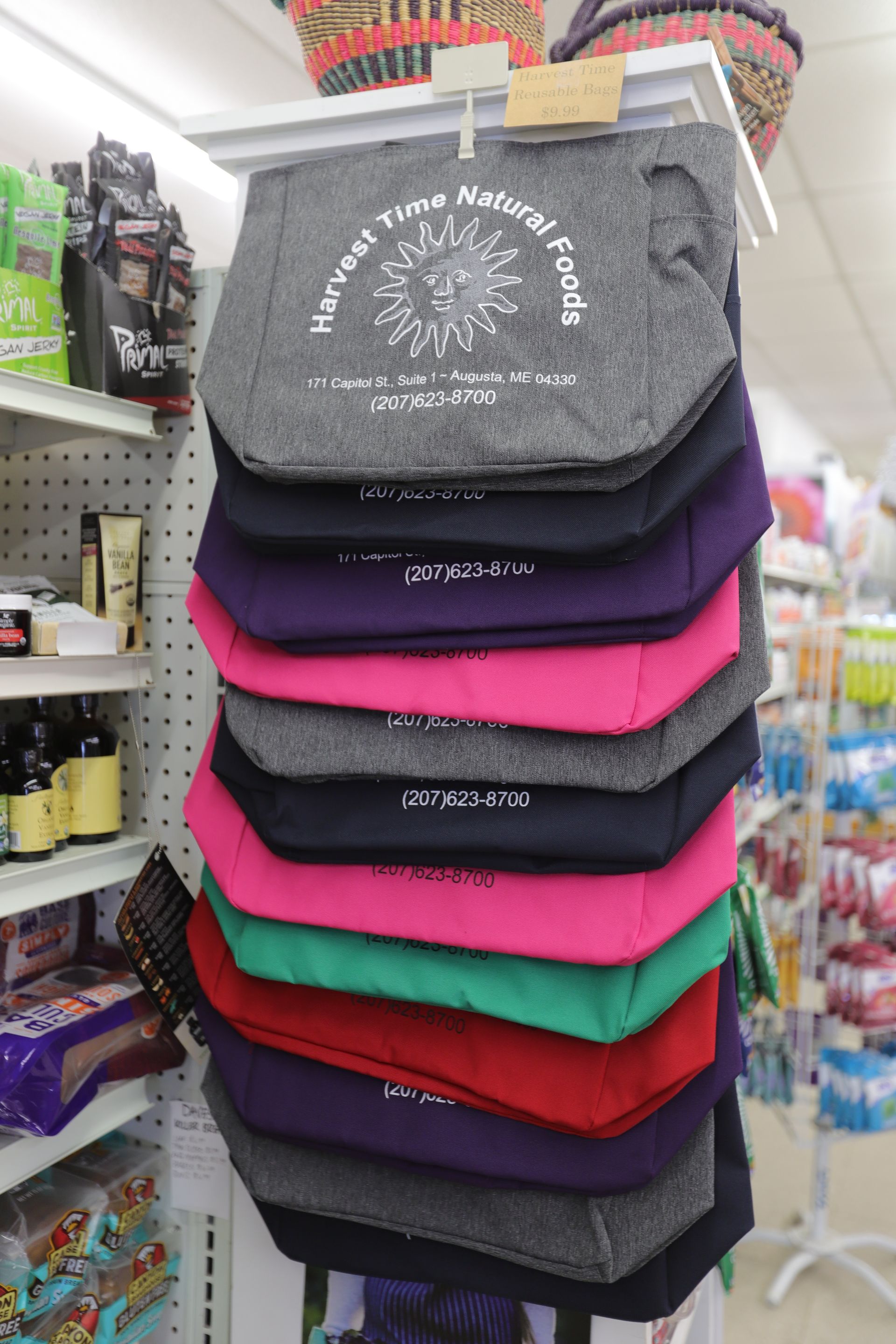 A vertical stack of branded reusable tote bags in various colors, including grey, purple, pink, green, and red.