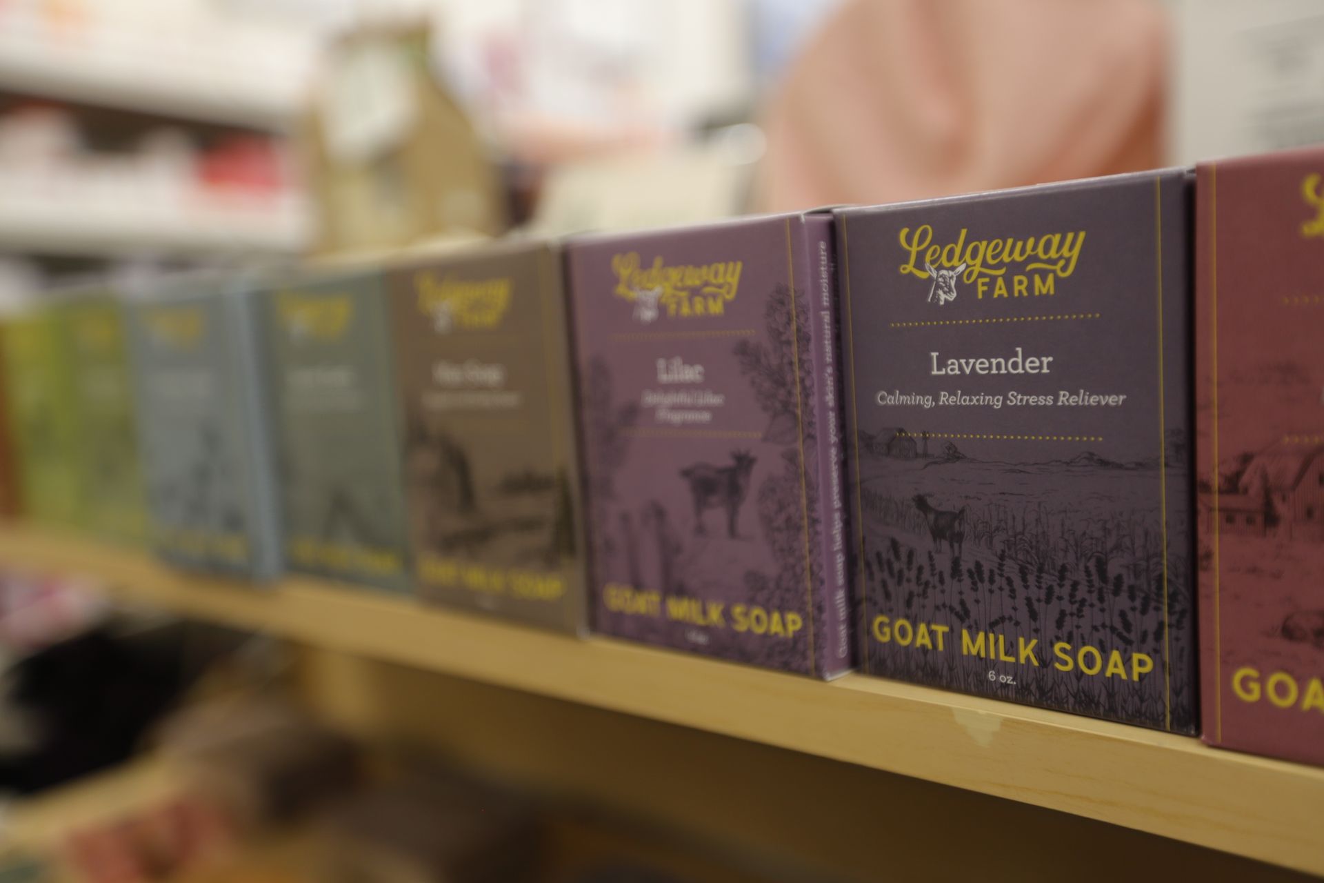 A row of Ledyeway Farm goat milk soap bars on a store shelf, featuring colors like lavender, purple, and brown.
