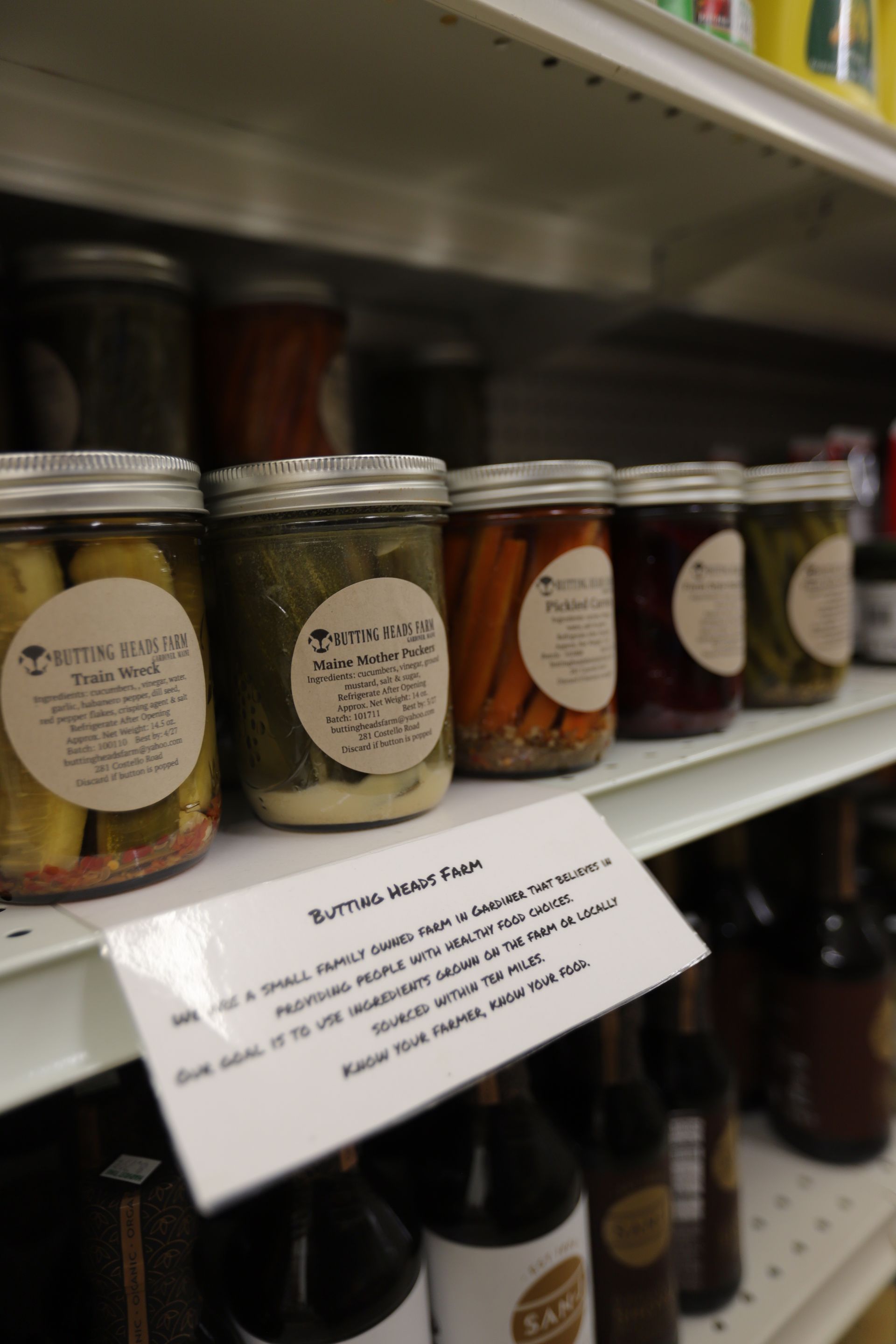 A store shelf displaying jars of pickles and vegetables with labels and a handwritten sign in front.