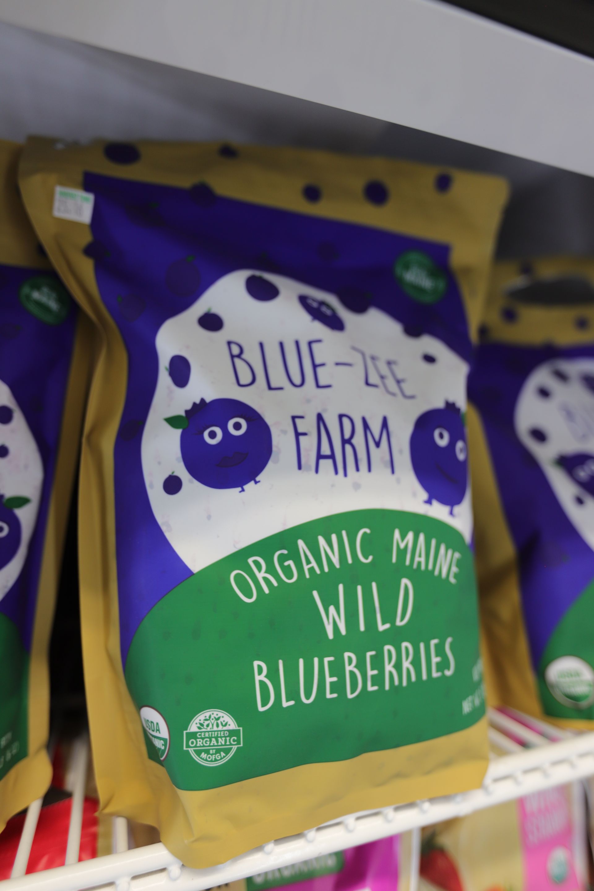 A bag of Blue-Zee Farm organic Maine wild blueberries sits on a store shelf, featuring cute cartoon blueberry graphics.