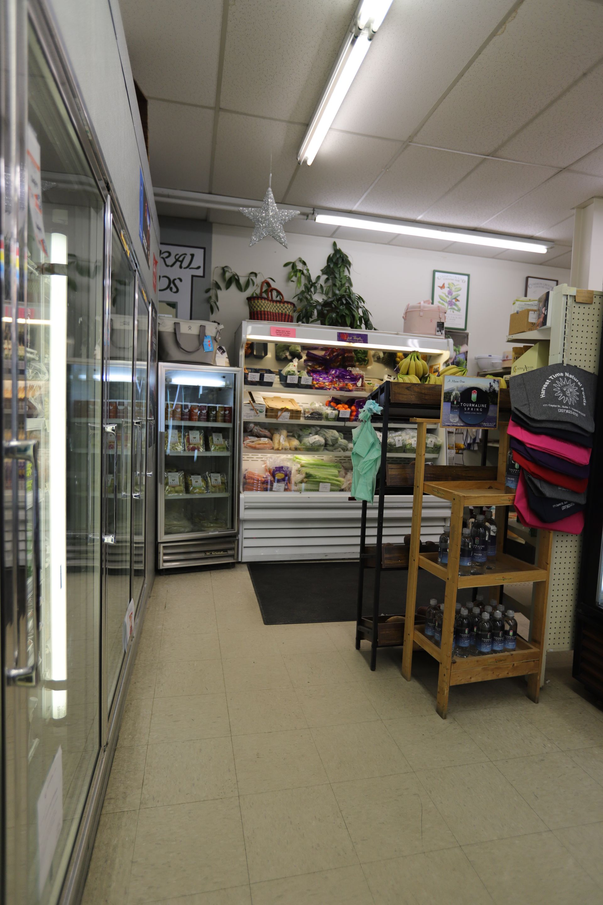 A small grocery store aisle with refrigerated display cases, a produce cooler, and a wooden display rack.