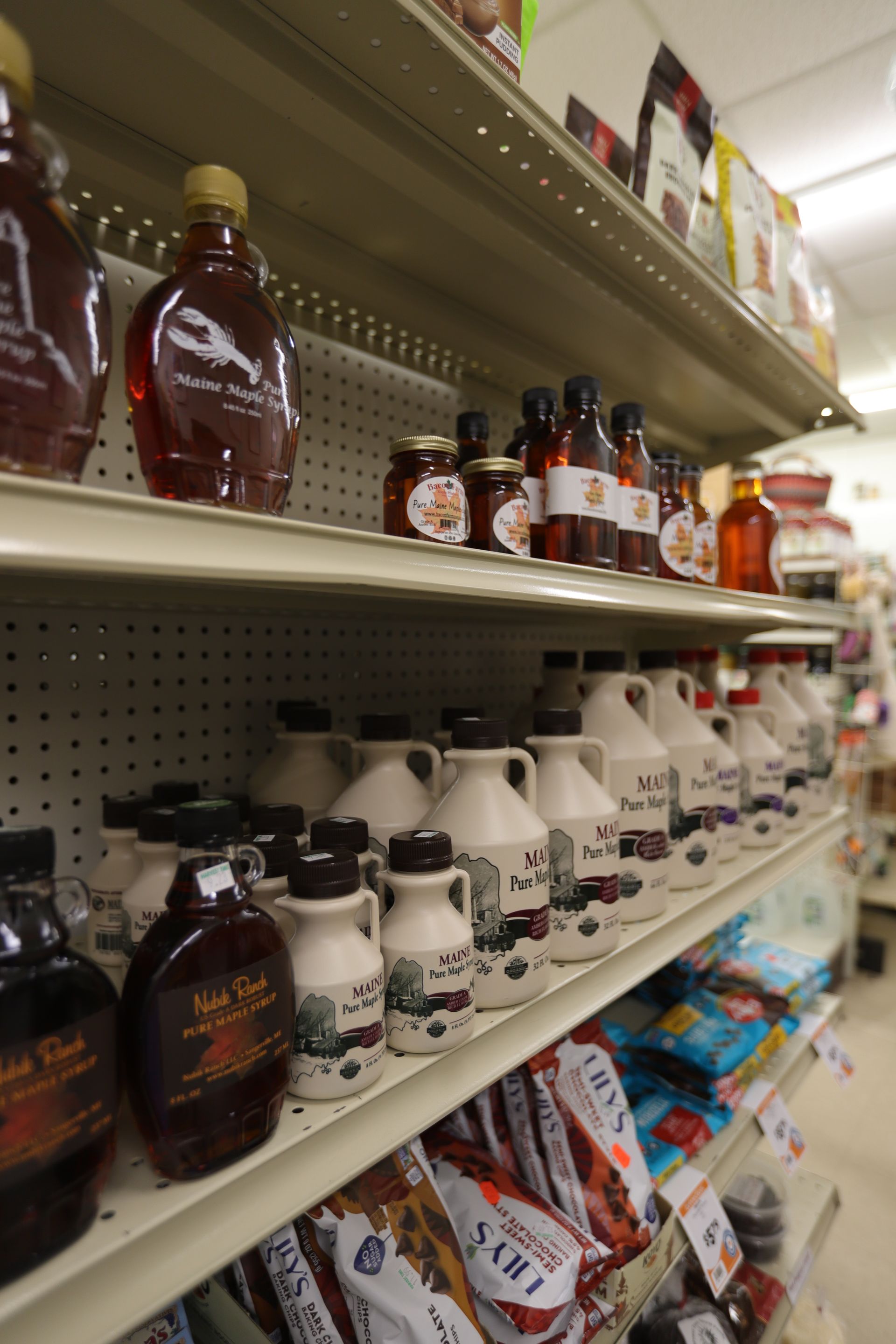 Shelves in a grocery store stocked with various glass and ceramic bottles of maple syrup and bagged goods.