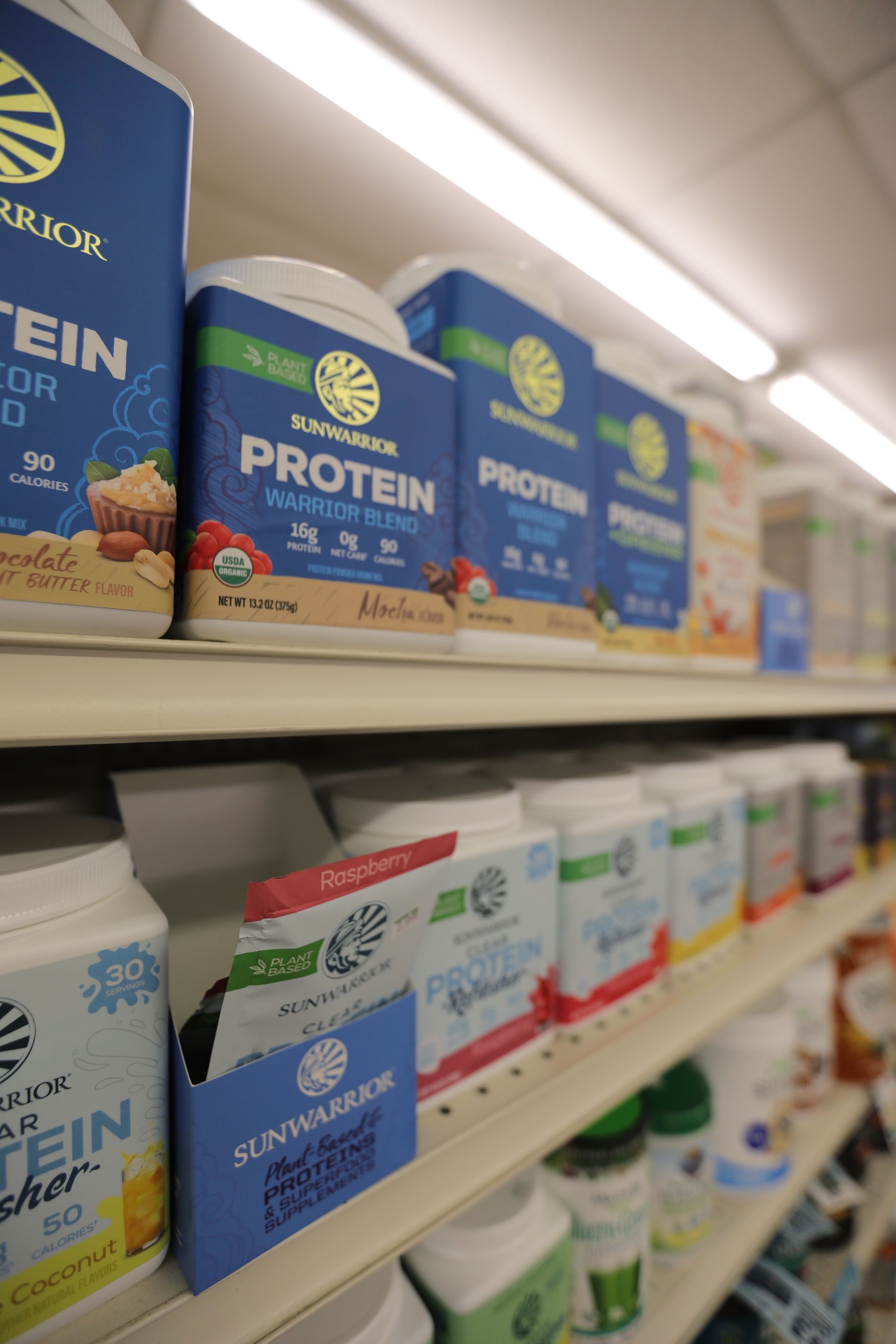 Shelves filled with various Sunwarrior protein powder containers and packets in a brightly lit retail store.