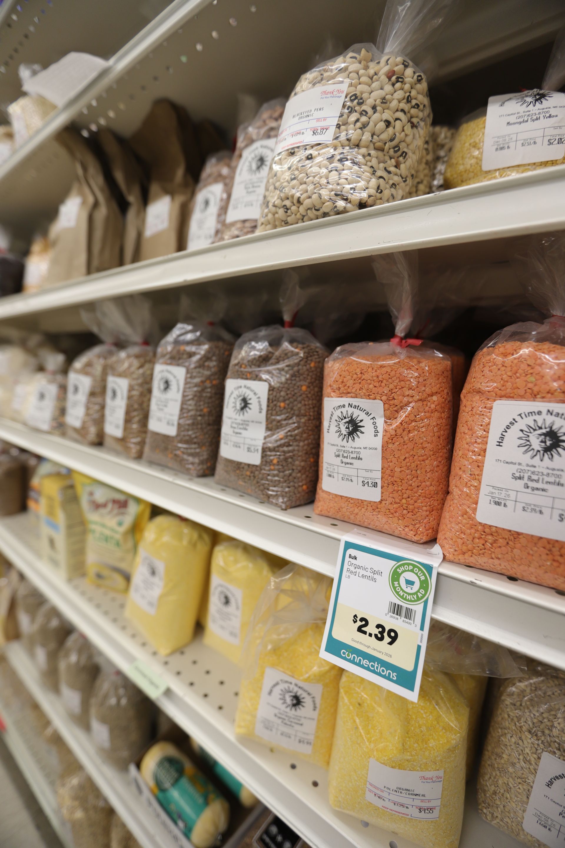 Shelves in a market stocked with various grains and legumes in clear plastic bags, including red lentils and yellow corn.