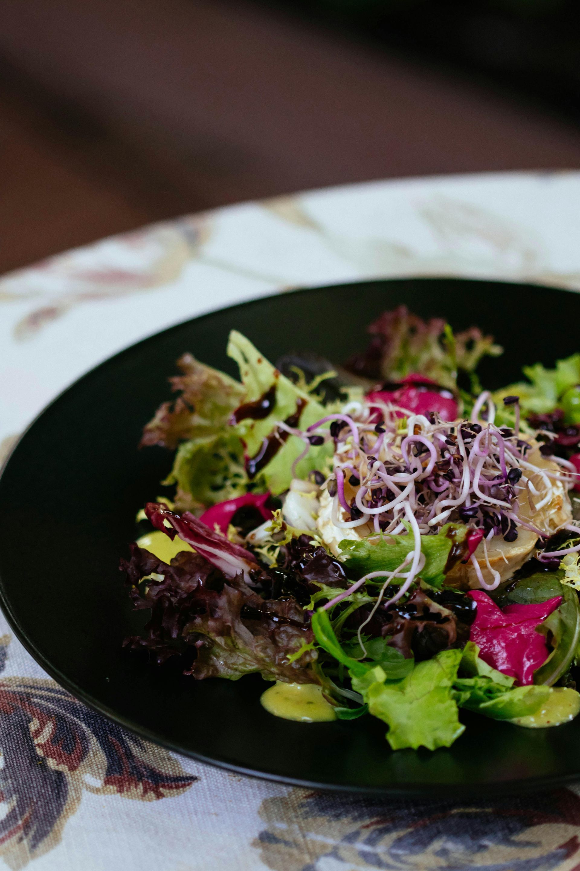 A salad with greens, sprouts, and dark balsamic glaze on a black plate, set against a floral tablecloth.