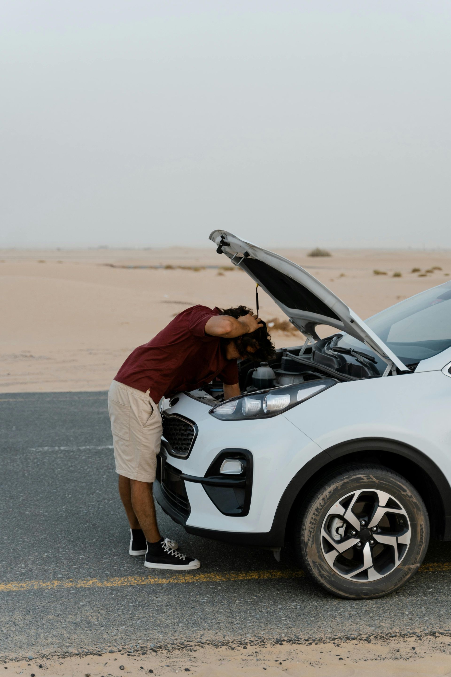 Person leaning into the open hood of a white car on a desert road