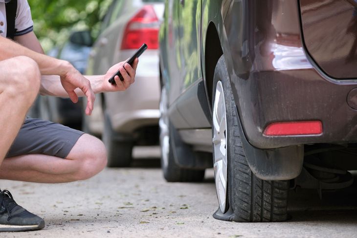 Person crouching beside a car with a flat rear tire, inspecting it with a phone on a driveway.