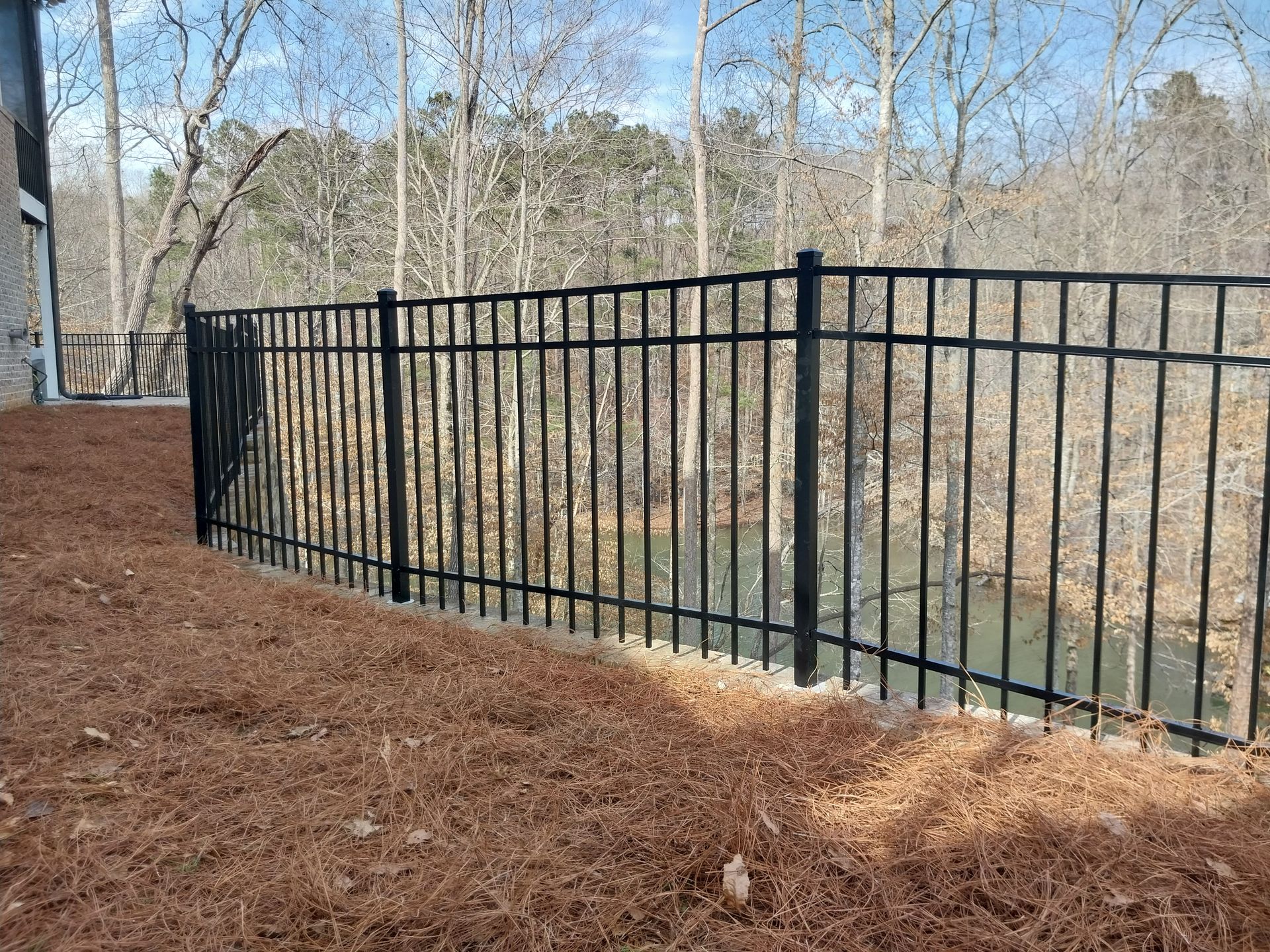 Black metal fence along a wooded slope with brown ground cover and bare trees