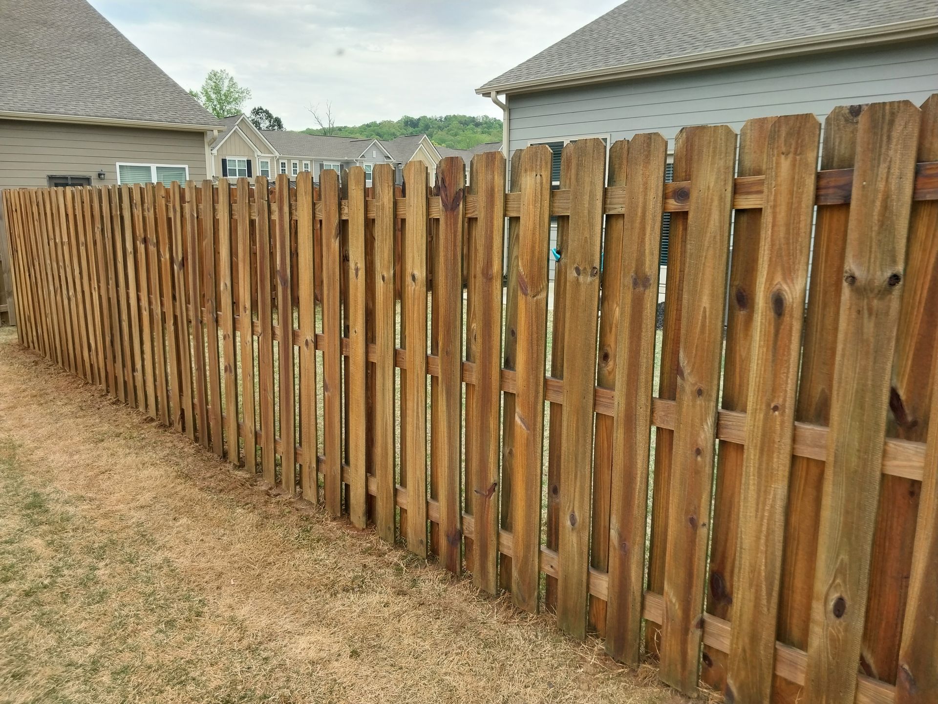 Weathered wooden privacy fence along a dry yard beside houses, with a cloudy sky overhead