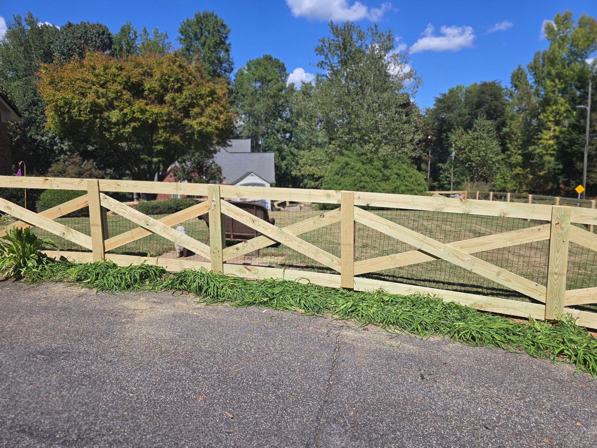 Wooden gate with diagonal braces along a gravel road, opening to a grassy field with trees and blue sky.