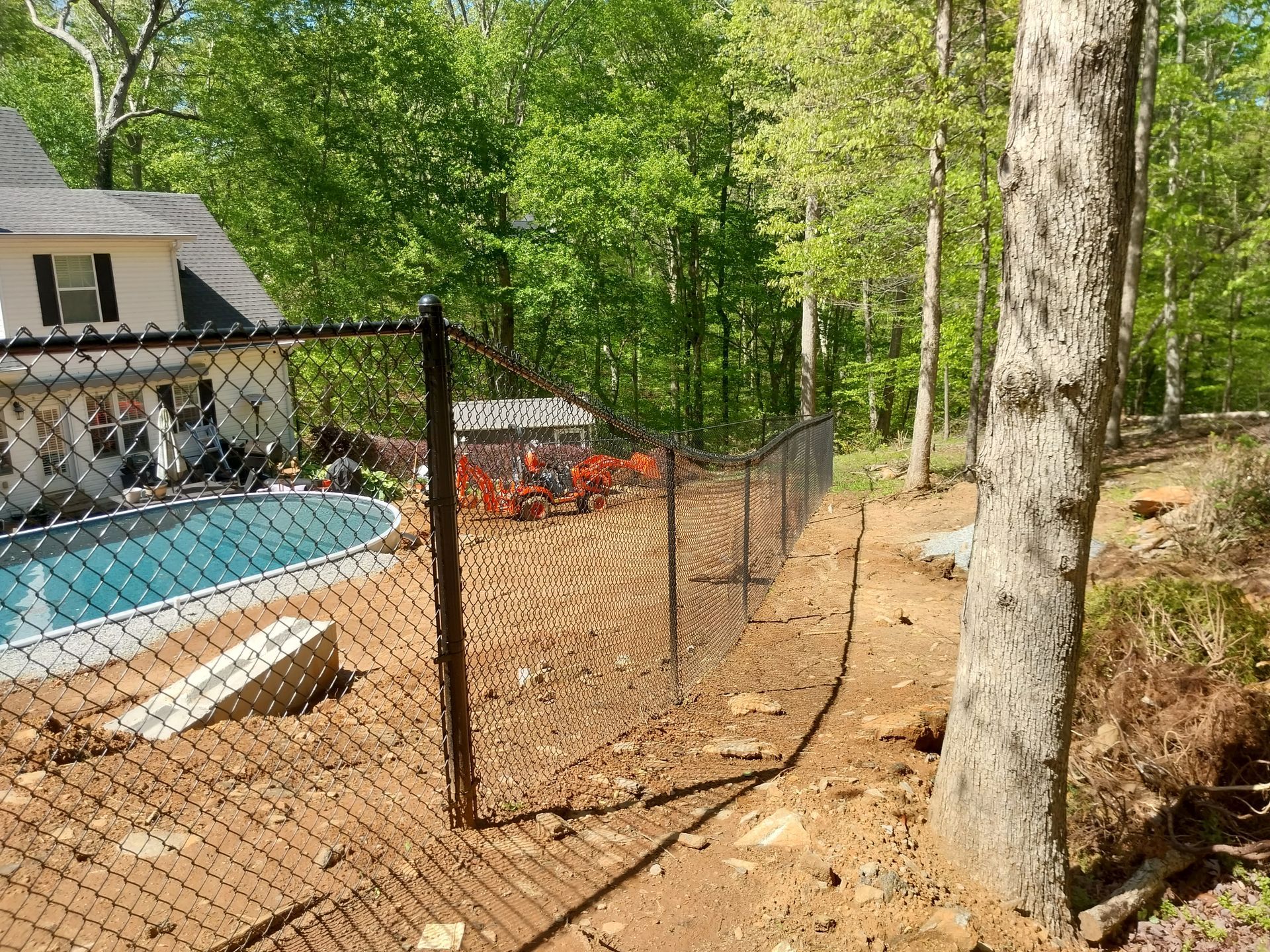 Chain-link fenced yard with pool, patio furniture, and trees in a wooded backyard