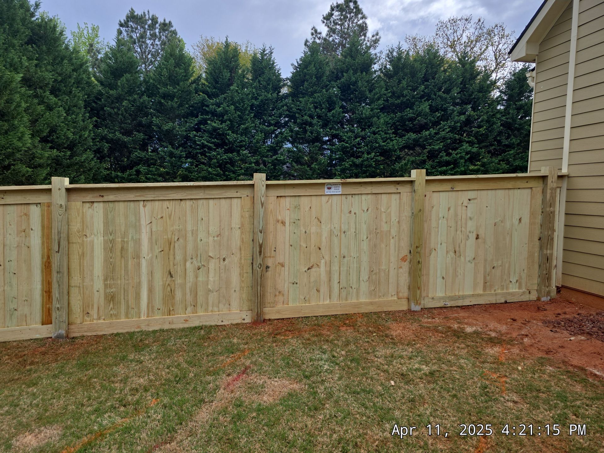 New wooden privacy fence in a backyard beside a beige house, with trees in the background.