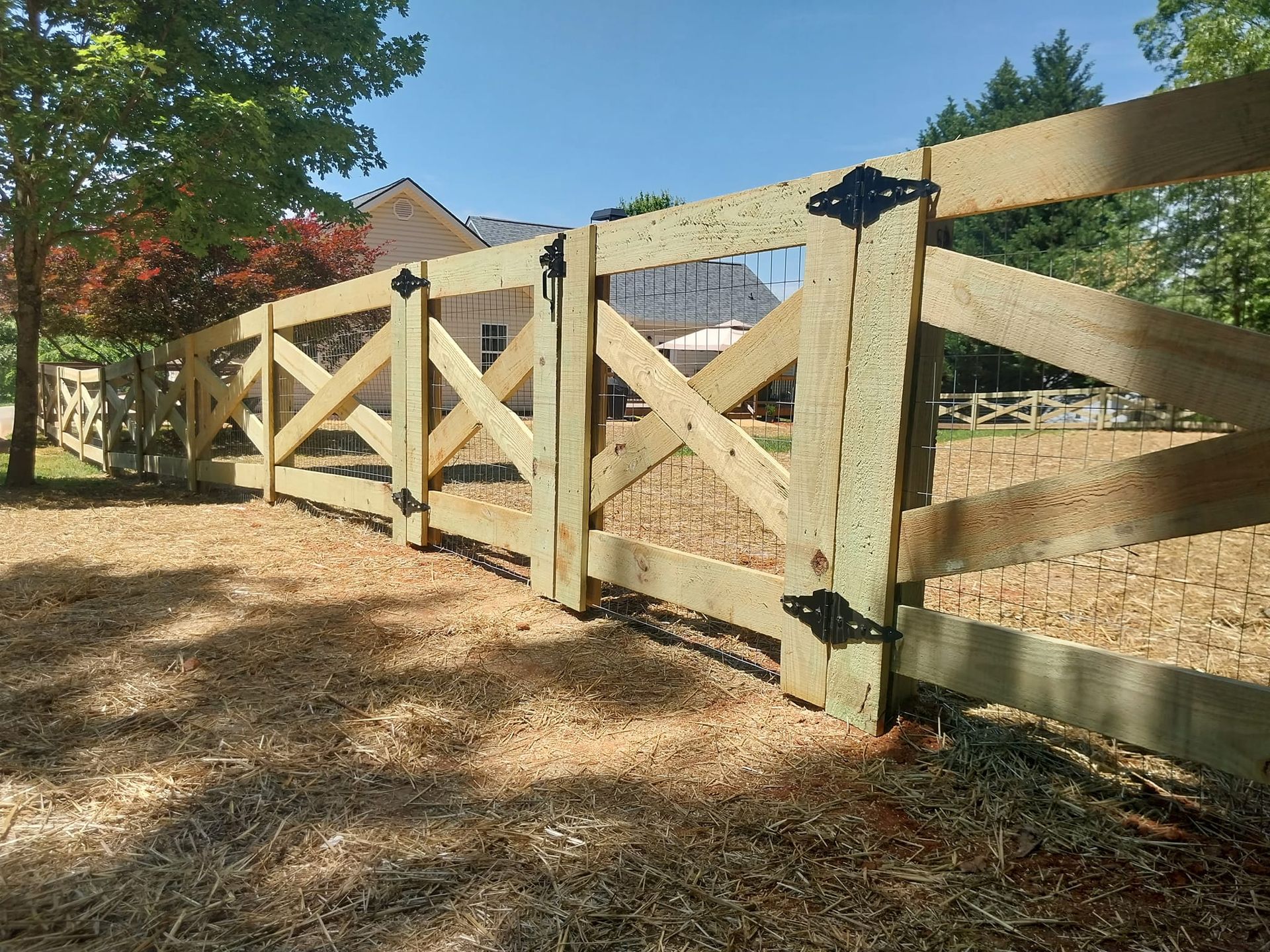 Wooden ranch-style gate with diagonal braces and black hardware along a tree-lined driveway in sunlight